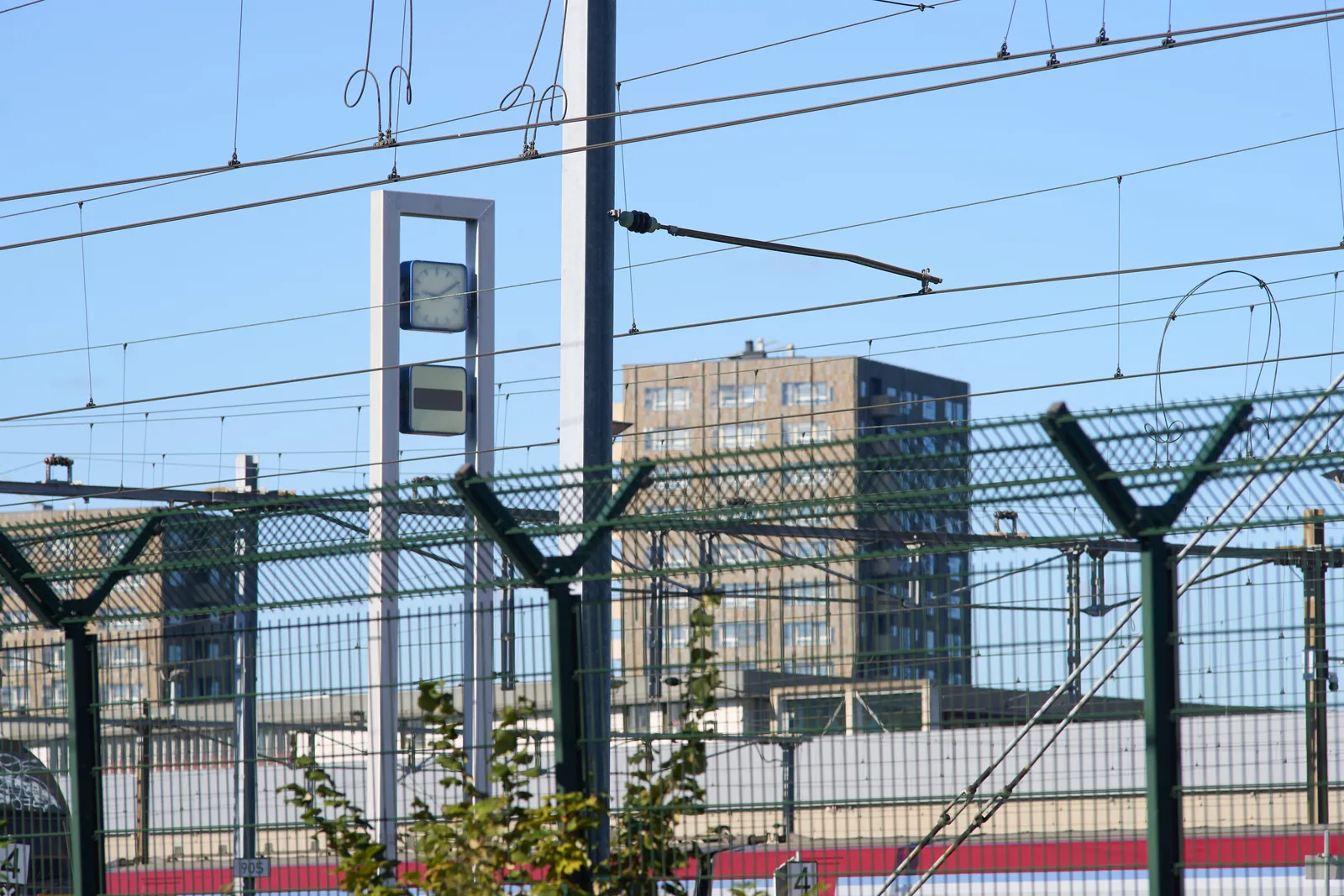 Fences, wires, poles, a large appartment building, and a clock, very high on two poles. It points at 10 past nine.