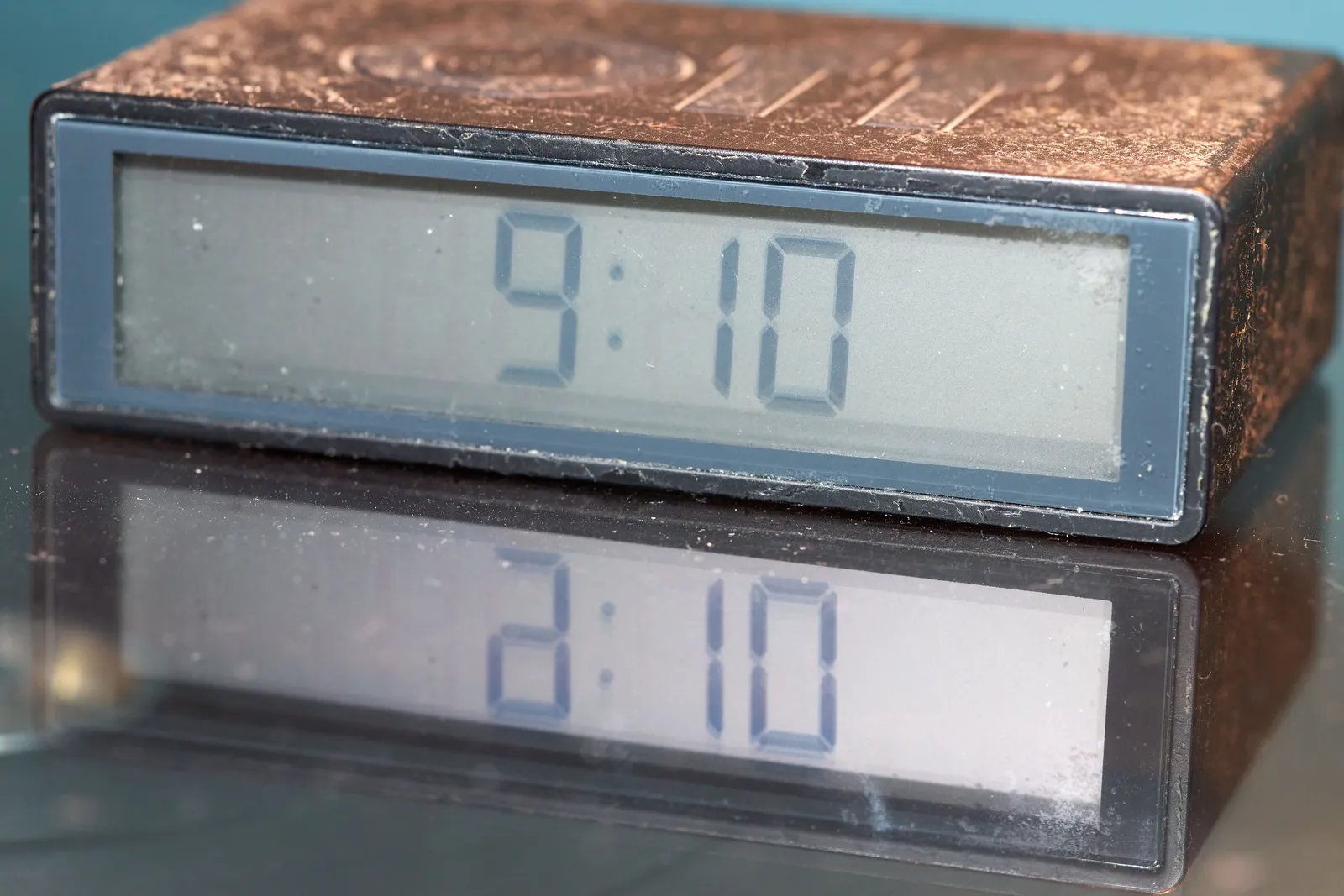 A digital alarm clock placed on something reflective. On the clock the time is displayed as 9:10