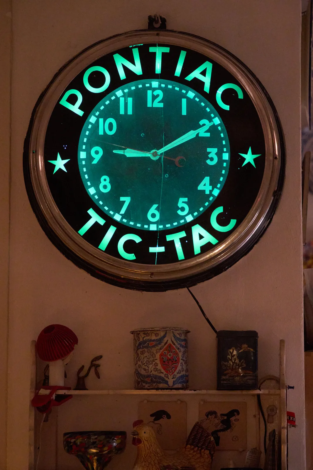 A neon lit wall clock with the words Pontiac Tic-Tac on it. It points at 10 minutes past 9. Below the clock is an assortment of little things on shelves.
