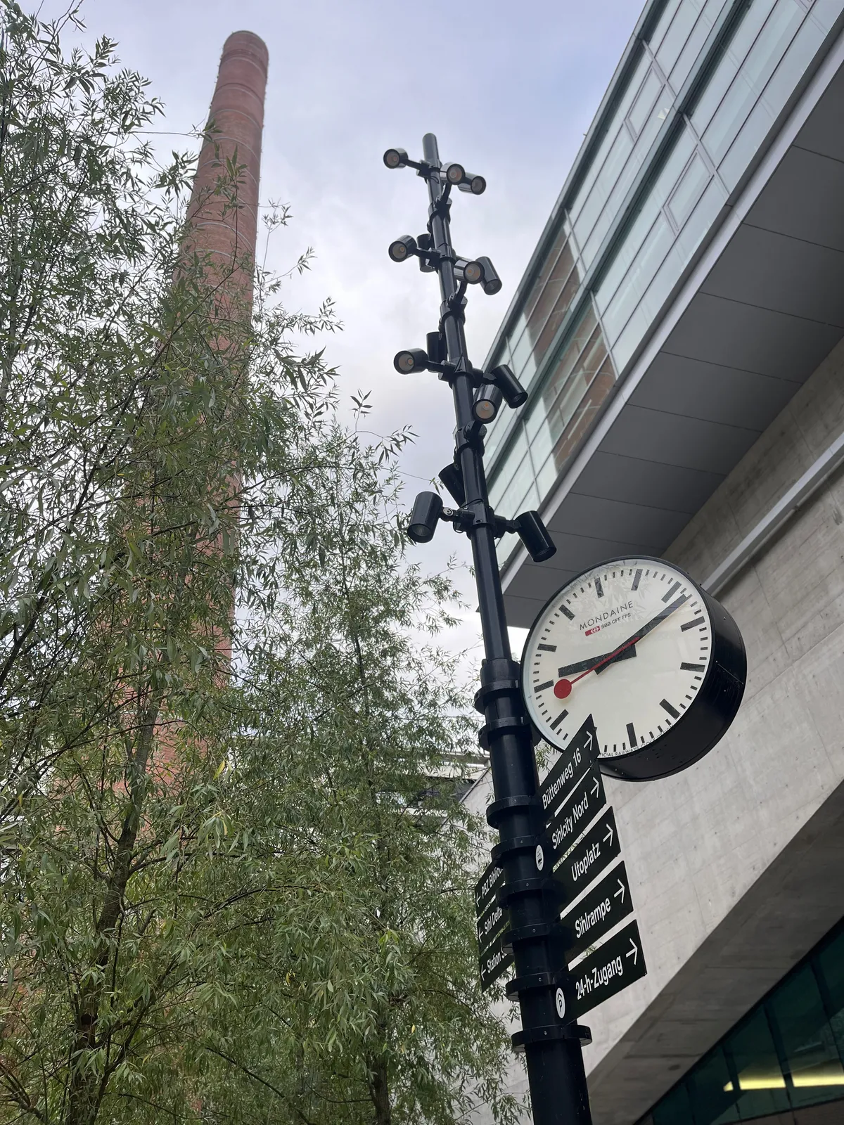In the background, behind a tree, there’s a very high chimney. Next to it there’s a clock, attached to a pole. On it it is 11 past 9.