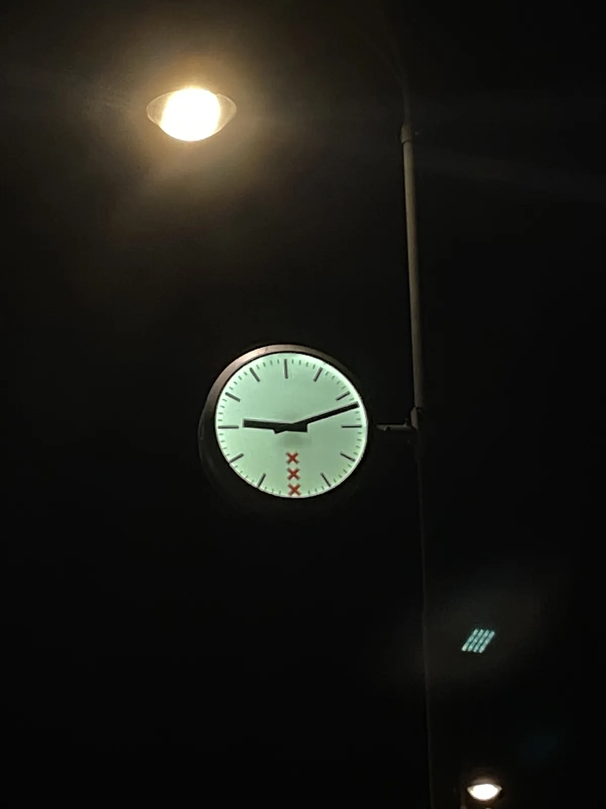 A municipal clock in Amsterdam at night. The clock is bright against the dark sky, just like the street lantern above it. The clock points at 12 past 9.