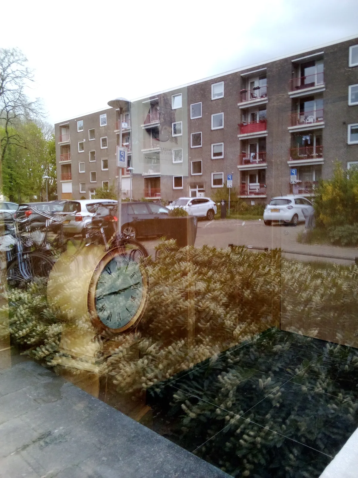 A brick appartment building is clearly visible, reflected in a large window. It is harder to look through the window, but a large clock can be seen inside. It points at 14 minutes past nine.
