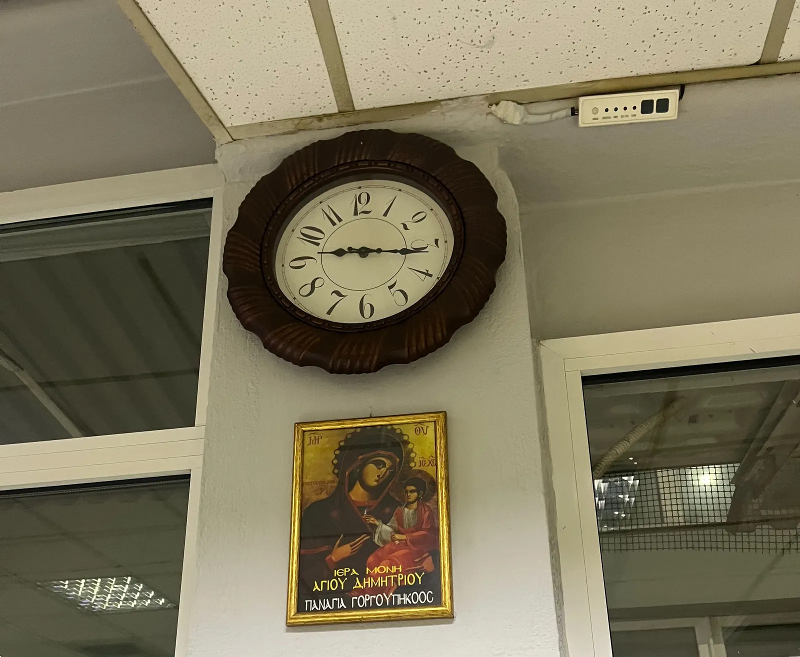 A large wall clock hangs over a Greek religious advertisement. The clock points at 16 minutes past nine.