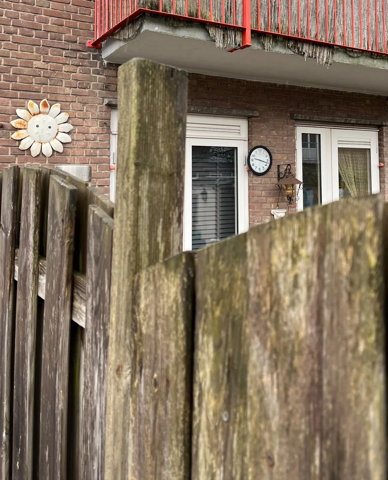 Looking over a fence into someones backyard. There’s a sun on the wall, and a clock. The clock points at 17 past nine.