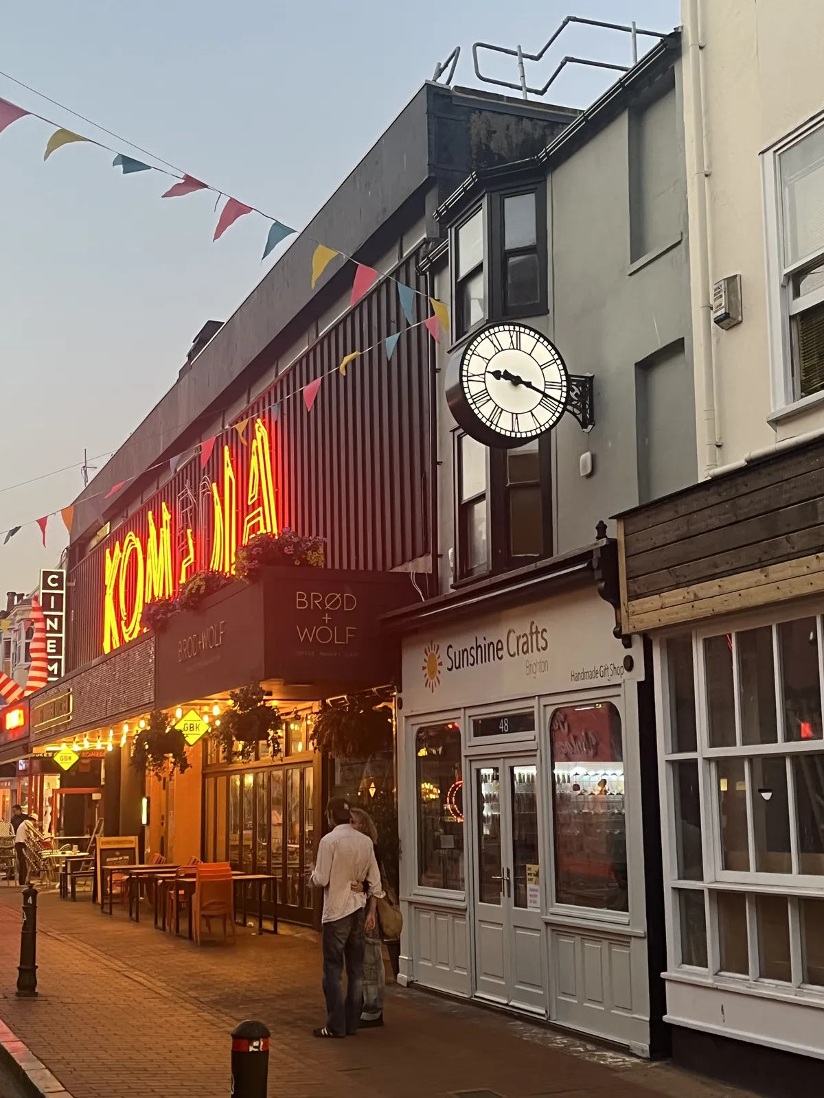 A clock attached to the facade of a small shop, next to a cinema with huge, brightly lit neon letters saying Komedia. The clock points at 18 minutes past nine.