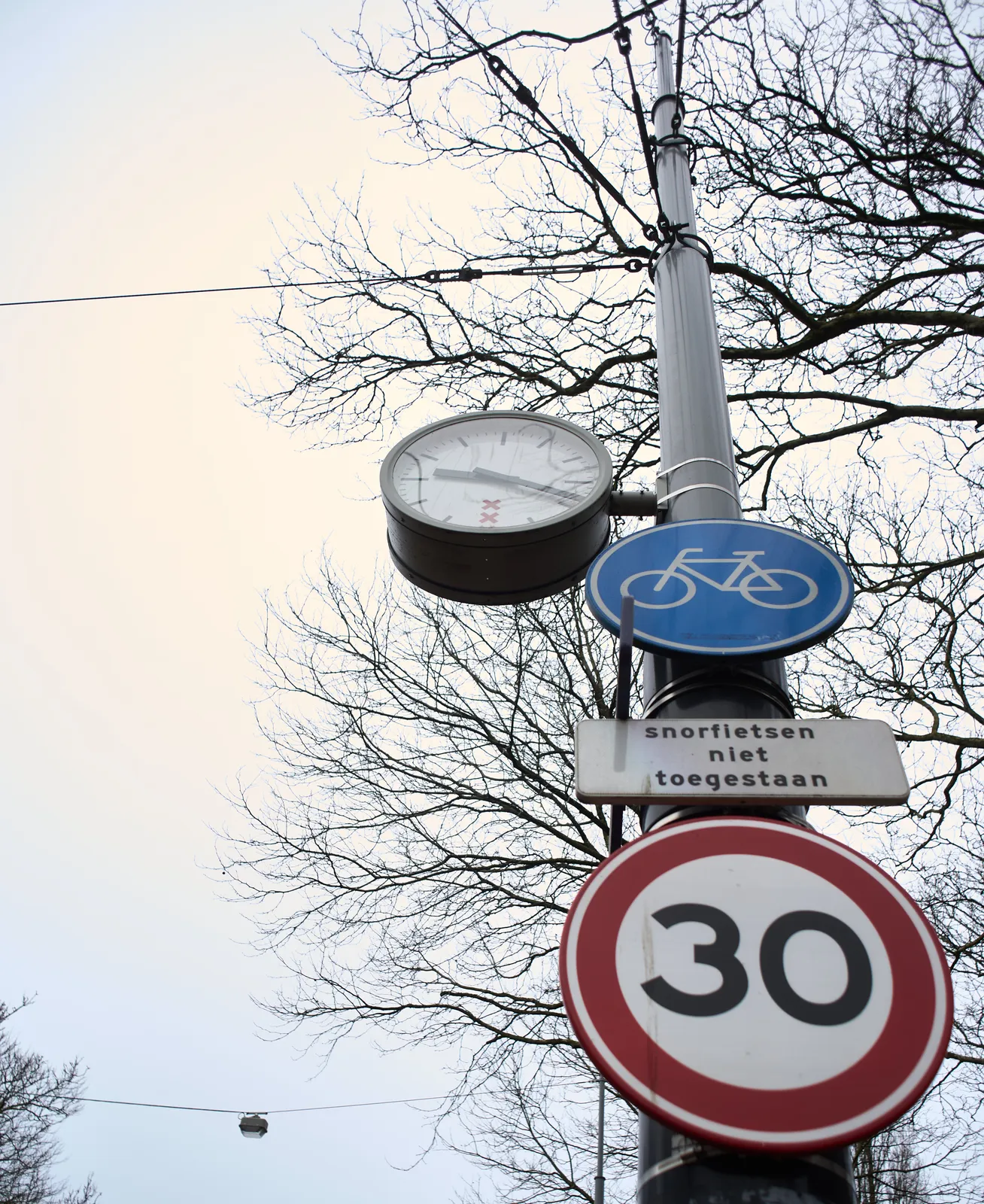 Looking up along a high pole with street signs, and a clock, attached to it. There’s a maximum speed of 30, it is not allowed to ride a moped, you are allowed to ride your bike though. The clock points at approximately 19 minutes past 9.
