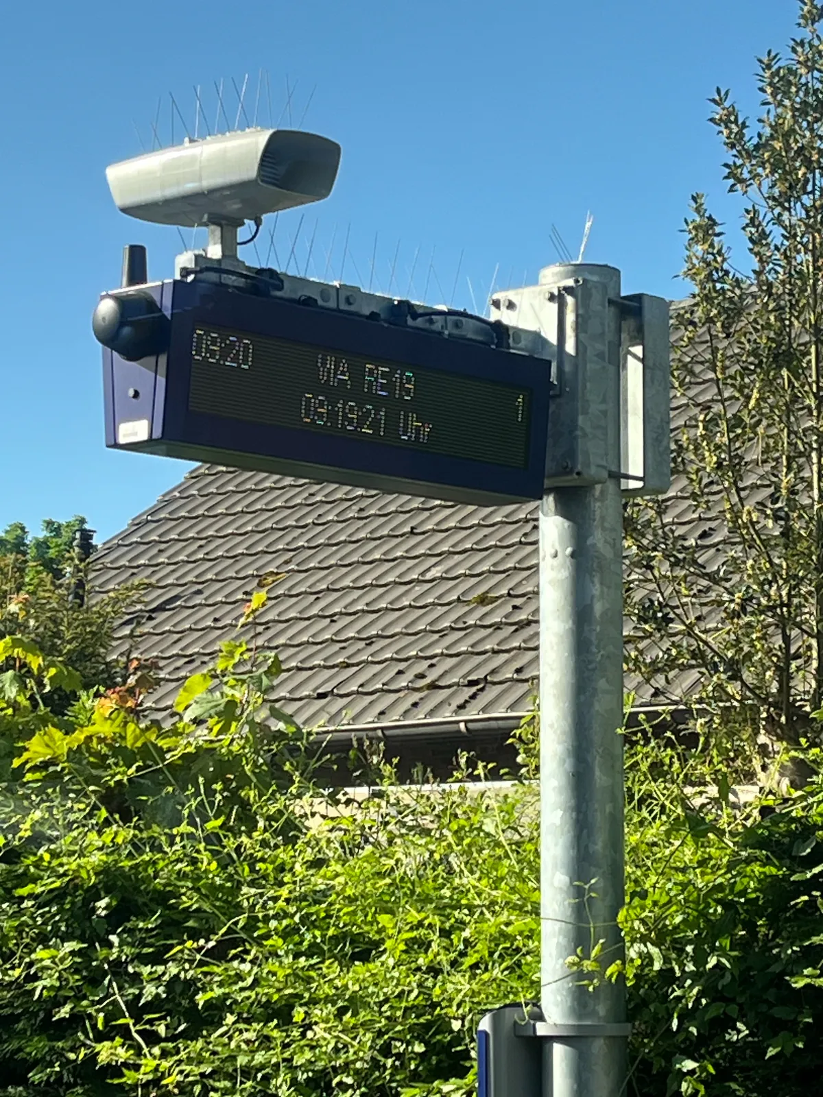 A digital display with a security camera on it. Both the camera and the display are filled with anti-bird spikes. On the display there are two timestamps. One says it is 09:19, the other says it is 09:20