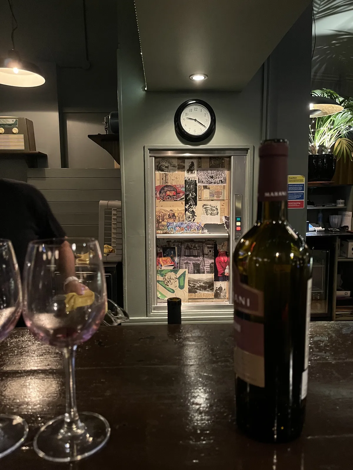 An empty wine glass and an empty wine bottle on a bar. Behind the bar, on the wall, is a wall clock which points at 20 past nine.