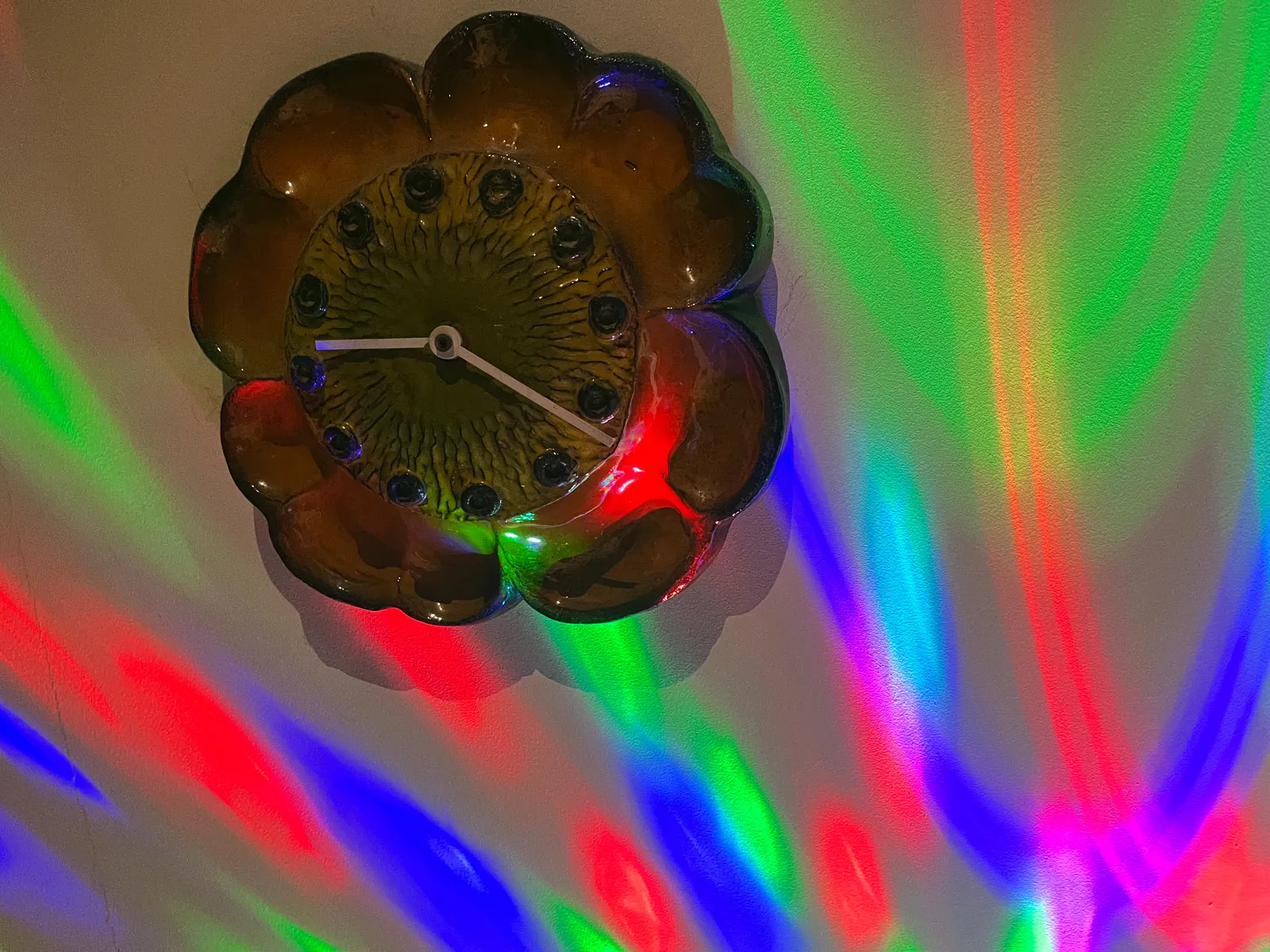 A cheramic wall clock. Bright coloured light shine onto the wall. The clock points at 21 minutes past 9