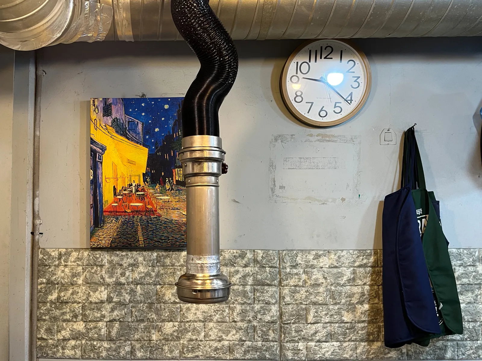 A very large silver ventilation shaft in the top of a picture. A smaller shaft is hanging from the ceiling. There are a few things hanging on the wall. A painting by Vincent Van Gogh. An apron. And a clock that points at 21 minutes past 9.