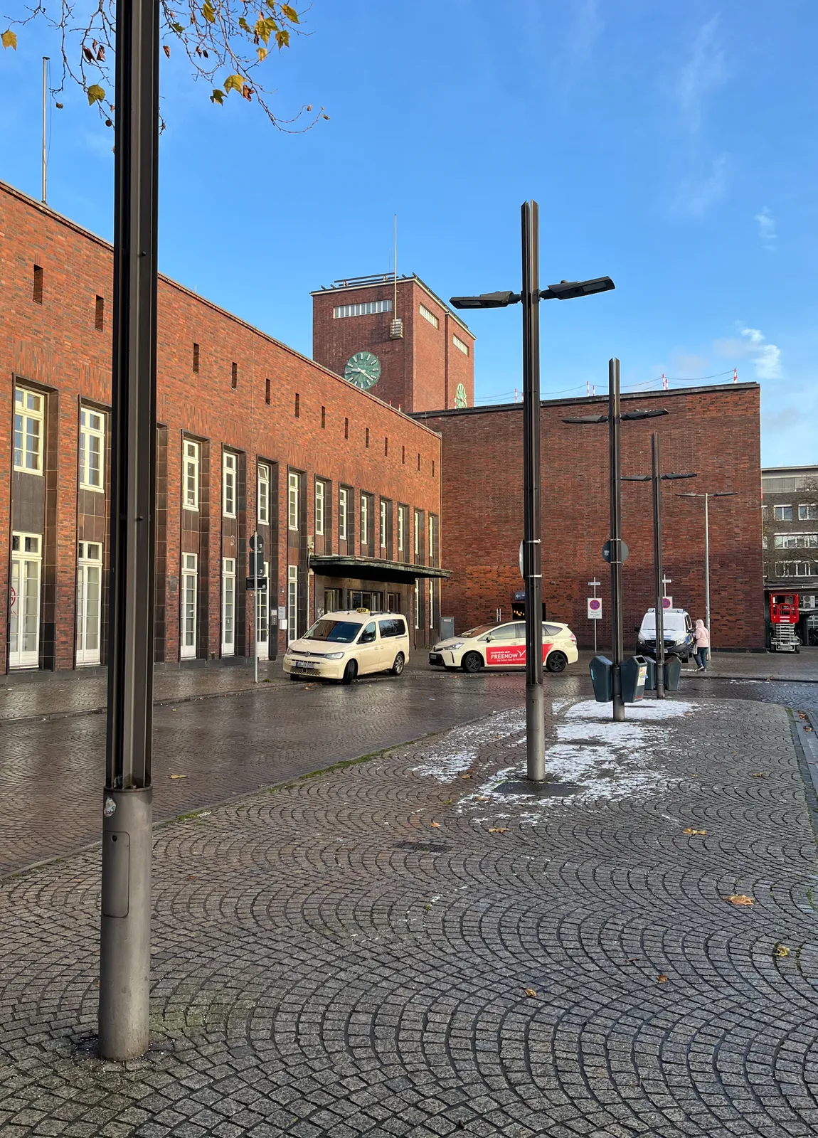 A monumental brick building, two white taxis, and a few cross-shaped lamp posts. There’s a clock on the building that points at 21 minutes past 9.