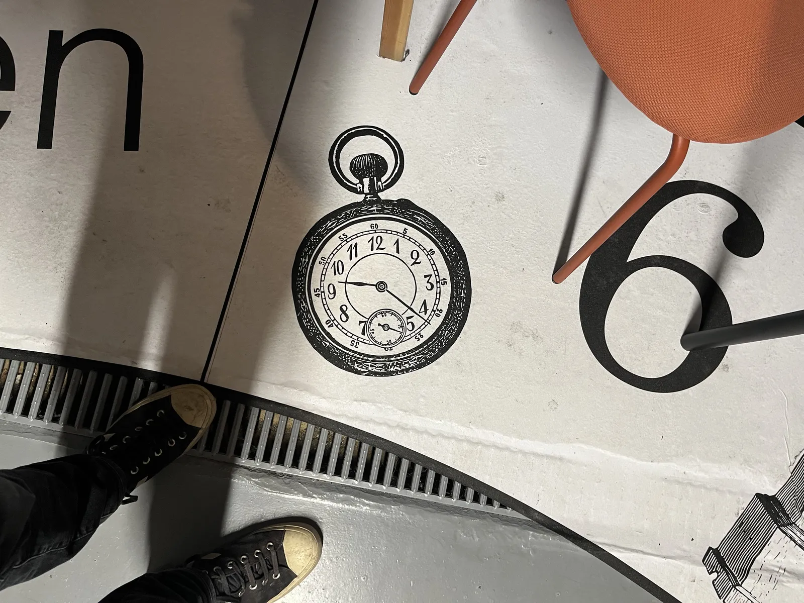 Looking down at the feet of the photographer who stands on a floor with an image of a pocket watch printed on it. This watch points at 22 minutes past nine.