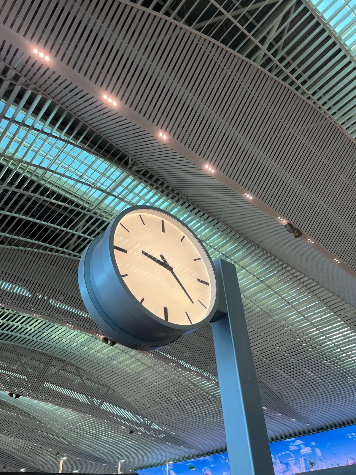 A minimalist clock, attached to a pole in a very large, very high hall. The clock points at 22 minutes past 9.
