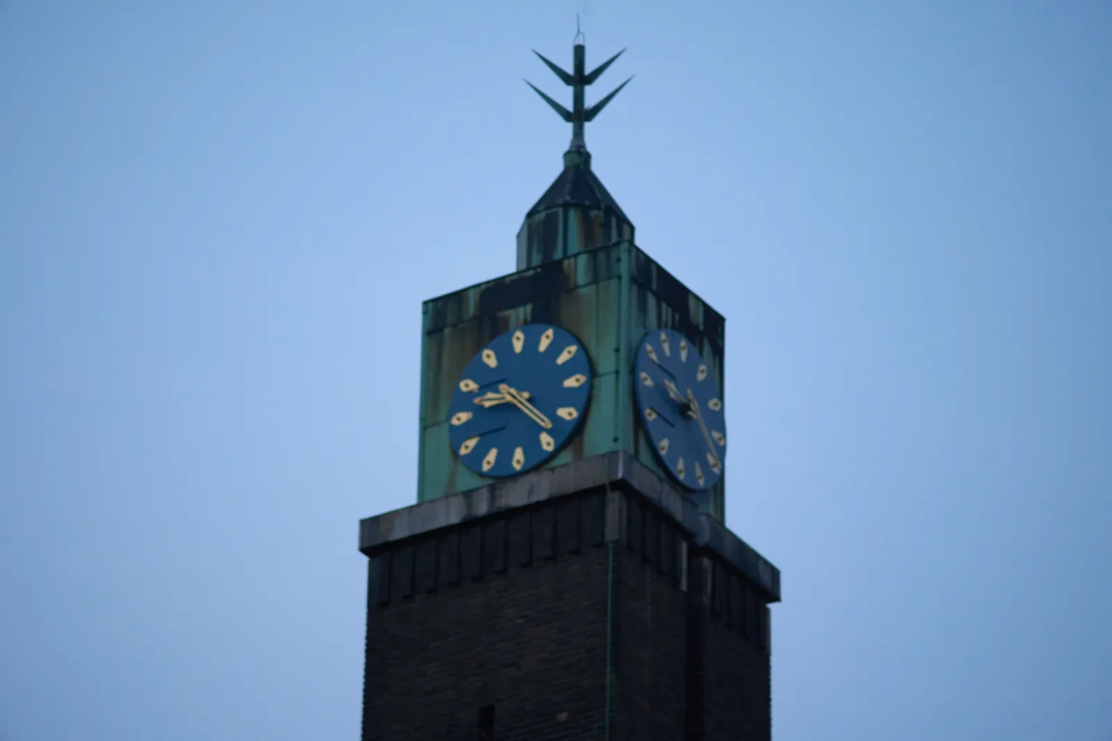 The top of a 20th century church tower. The base is made of bricks. The top is made from coroded copper, so it’s blue, with some brown stains. On the very top there’s a pin with some very pointy spikes. On both visible sides there’s a clock. On the side that’s best visible, the clock points at 23 minutes past 9.