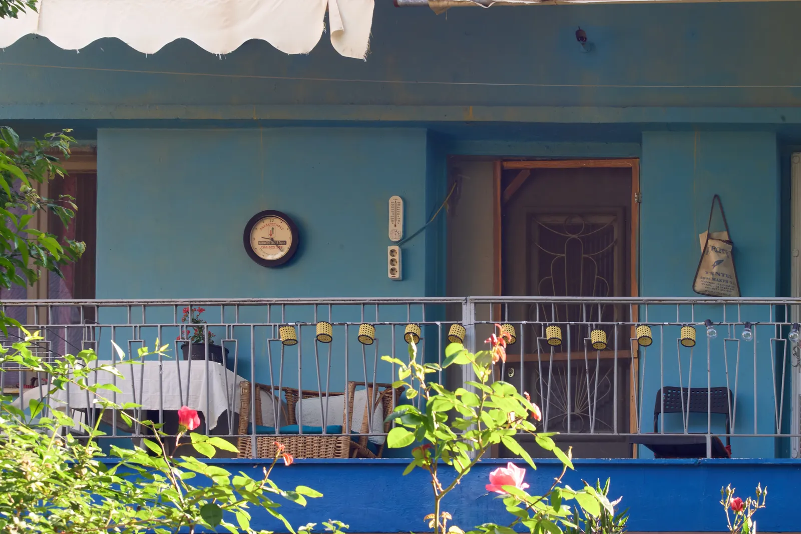 Peeking past some trees at the balcony of a blue building. On the wall there’s a clock that points at 24 minutes past nine.