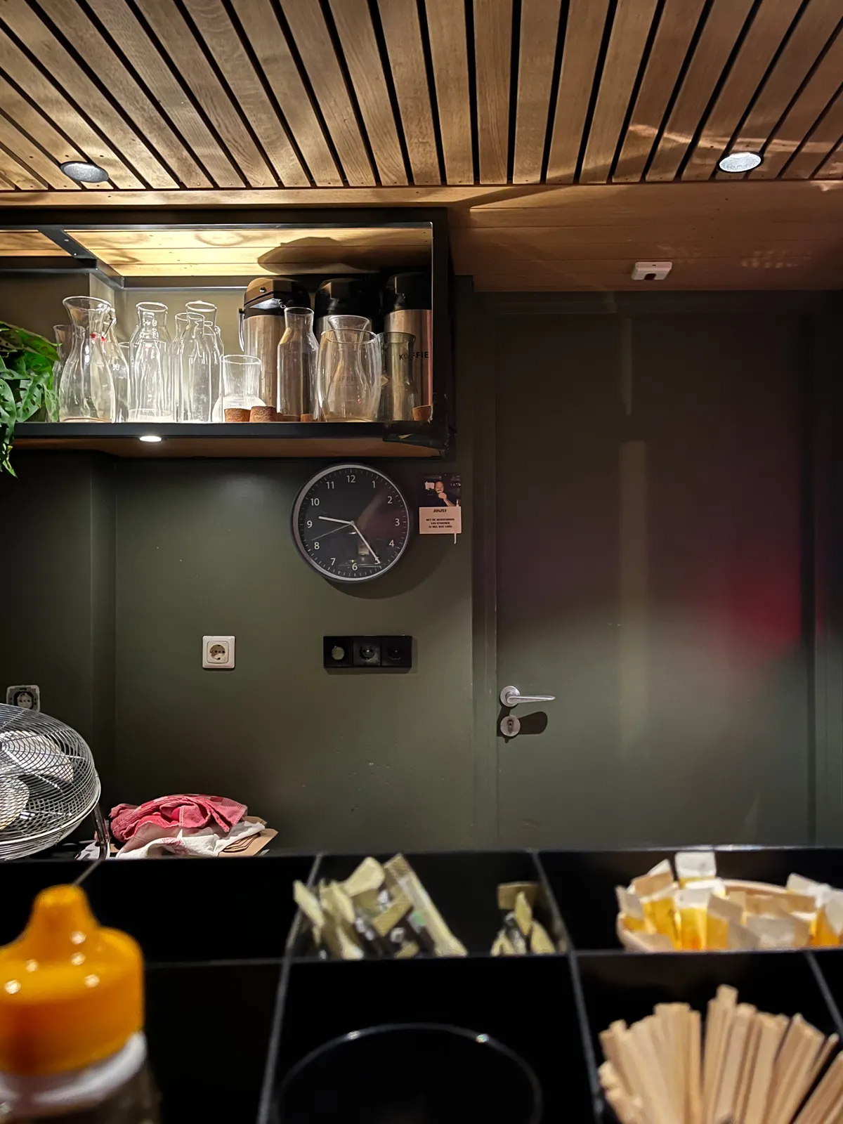 Looking over a bar, to the wall, and the wooden ceiling behind it. On the bar there are bags of sugar, and wooden spoons to stir your coffee. Behind the bar there’s a door, and a cupboard with glass carafes. There’s also a clock that points at 24 minutes past 9.