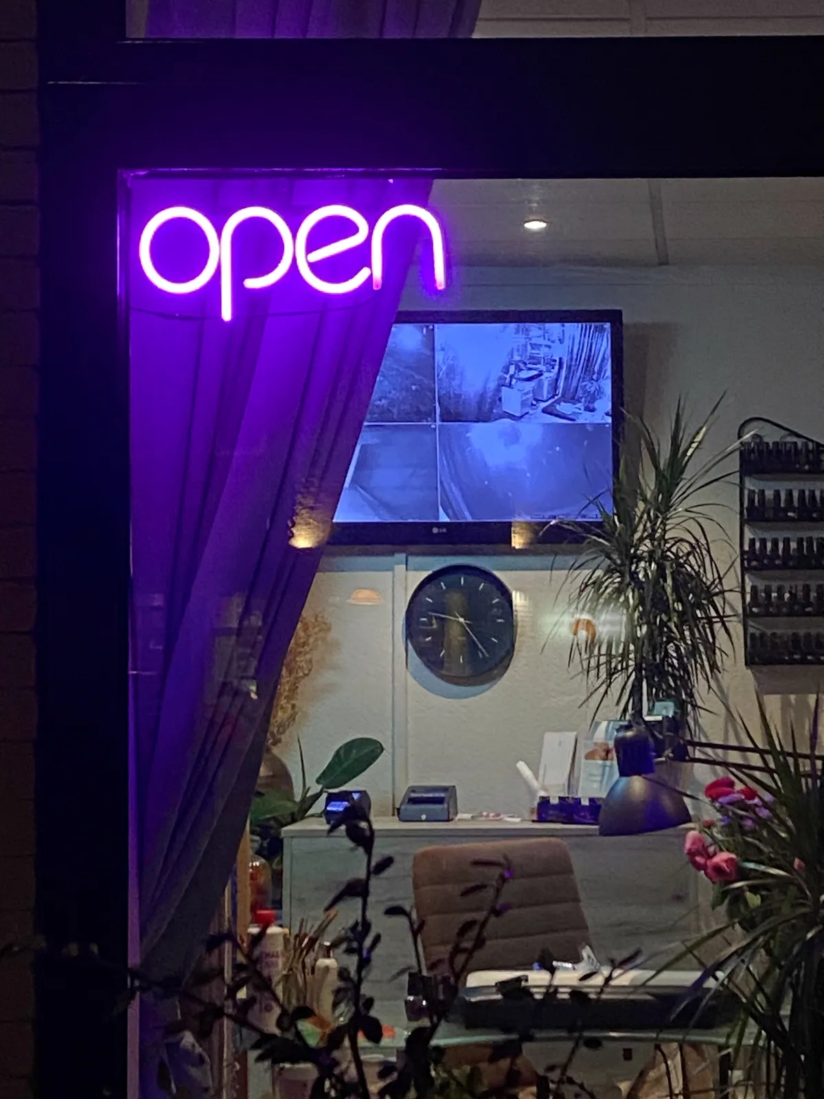 Purple neon letters say Open. Behind the sign some curtains, a chair and a clock. On the clock it’s 24 past 9.
