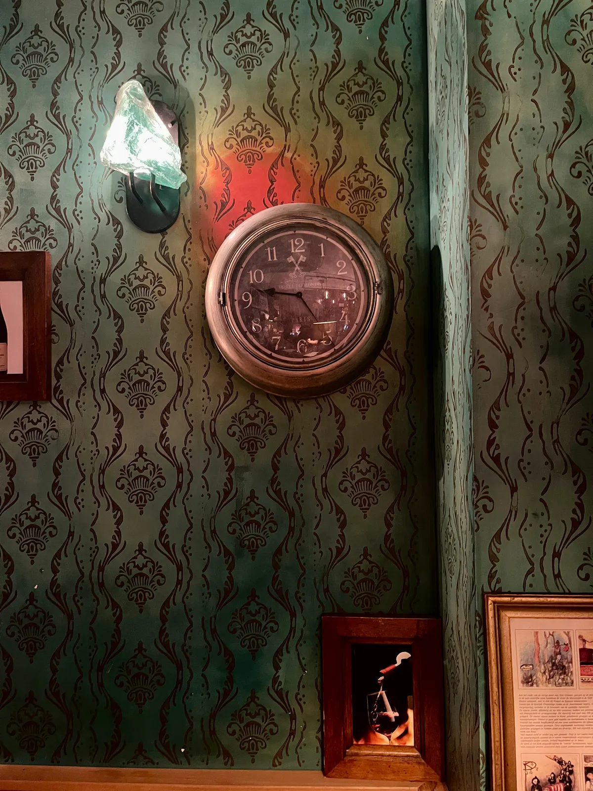 A wall with all kinds of pictures, lamps, and a clock. The clock points at 25 past 9.