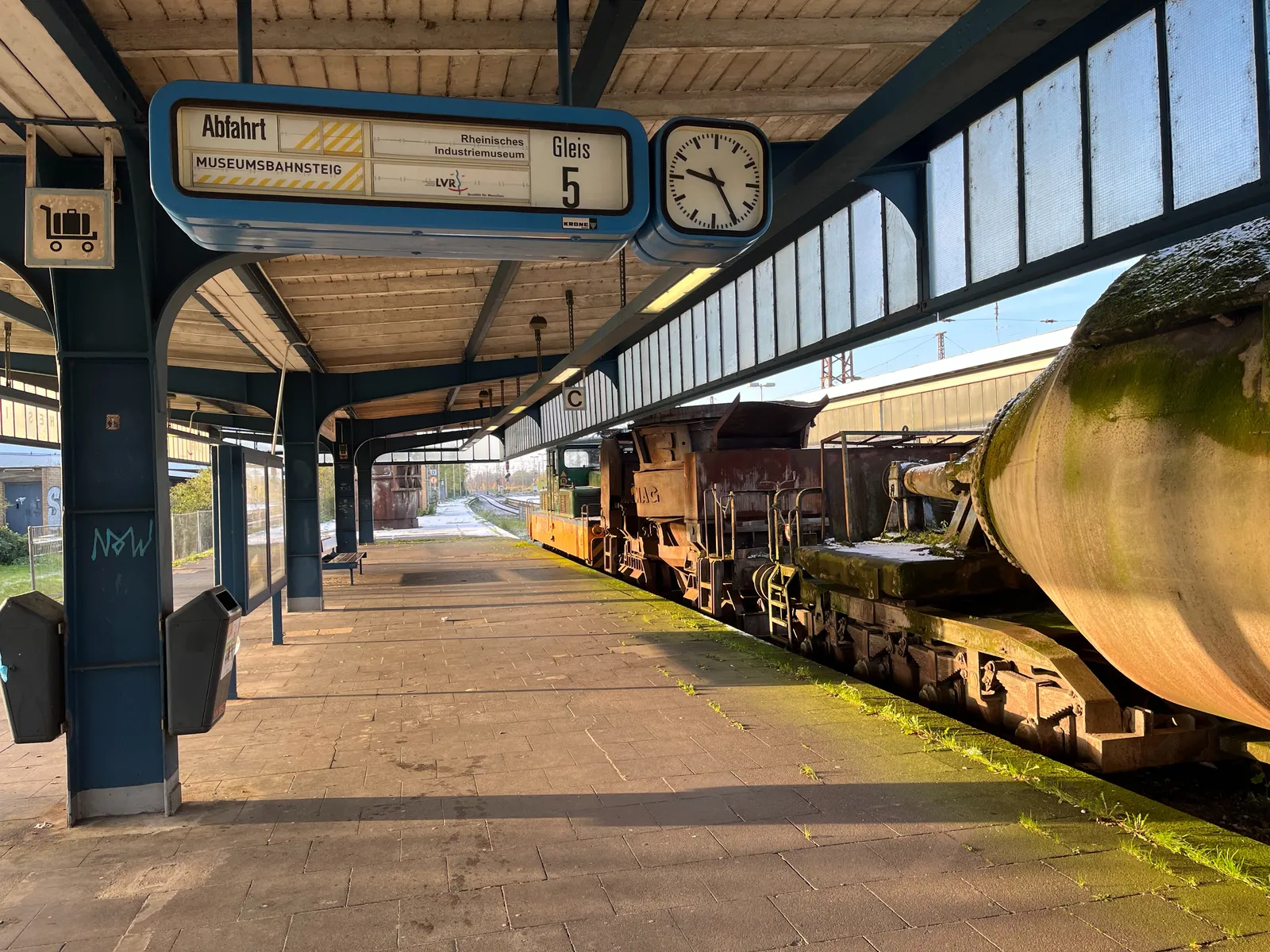 A mechanical display in a deserted train platform. The display says, in German, that this is the Museum Platform from the Reinisches Industriemuseum. The clock next to the display points at 25 past 9. There’s an impressive metal, very industrial train parked at the station. 