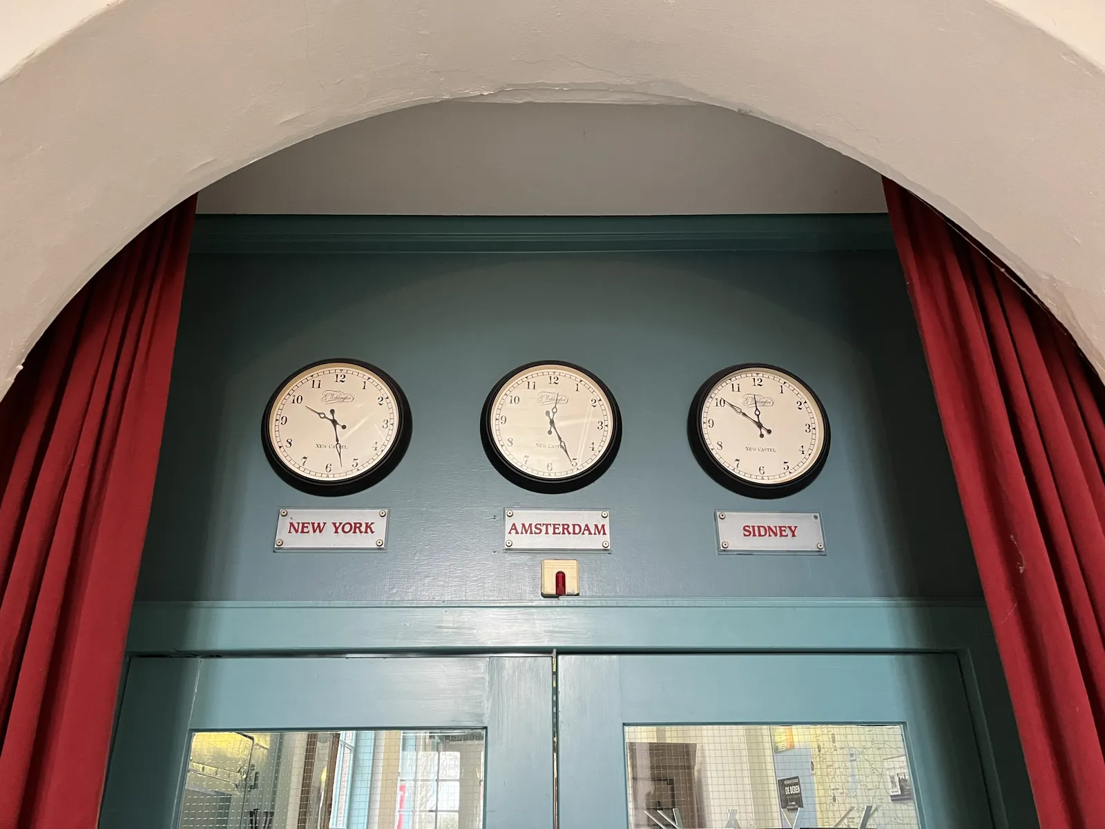 Behind an arc, there’s a blue door with open, red curtains. Above the door there are three clocks that all three show a different time. Below each clock is the name of a city: New York, Amsterdam and Sidney. On the first clock it is 27 minutes past 9, on the on in the middle it’s 25 minutes past 12, and on the one on the right it’s 9 minutes to twelve. 