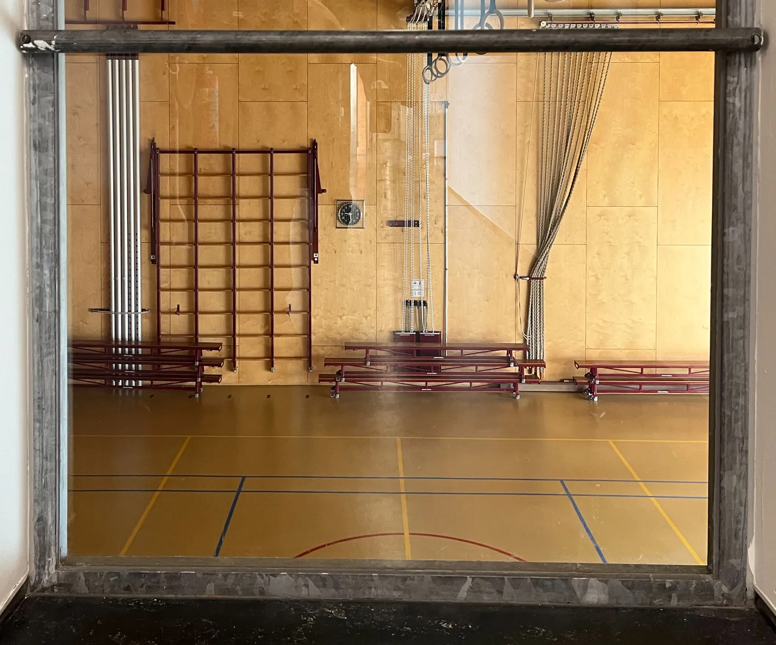 Peeking through a window into a gym, the ones that schools in the Netherlands have. There’s a clock on the wall in this gym, which points at 29 minutes past 9.