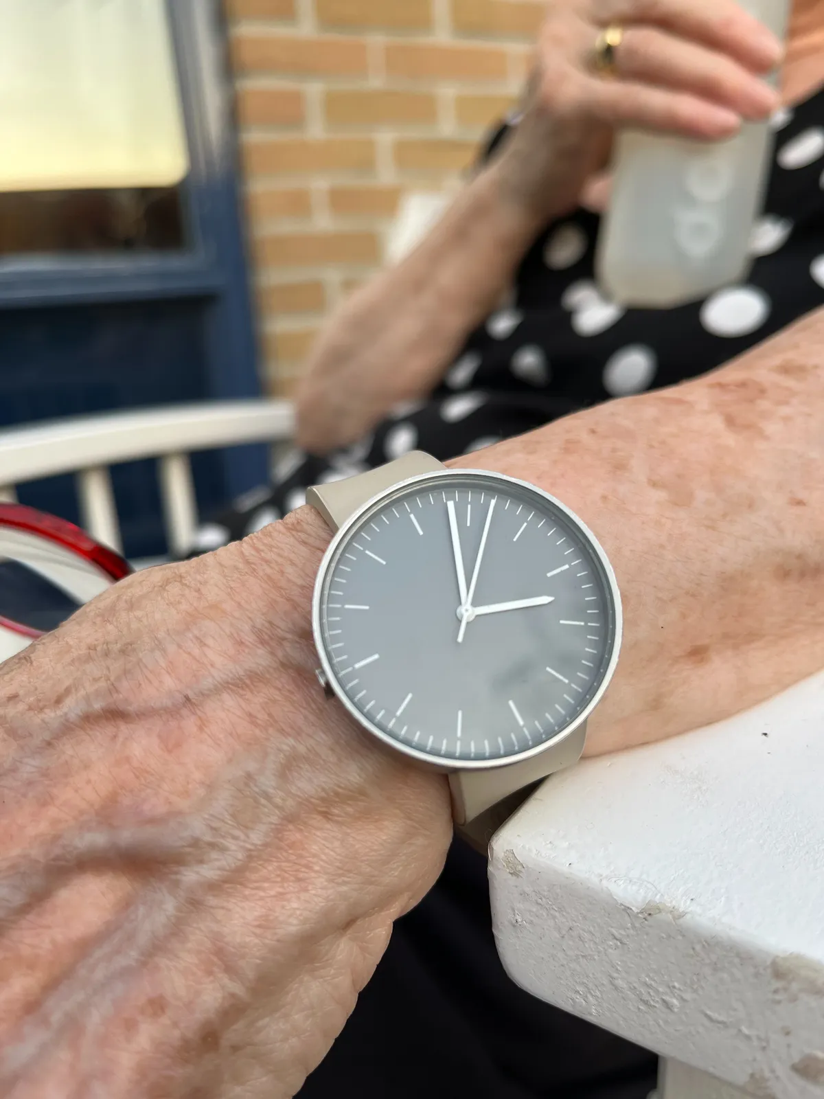 A gray, minimalist wrist watch on someone’s wrist. The watch is upside down, so it takes a bit of effort to see that it points at 27 minutes to 10.