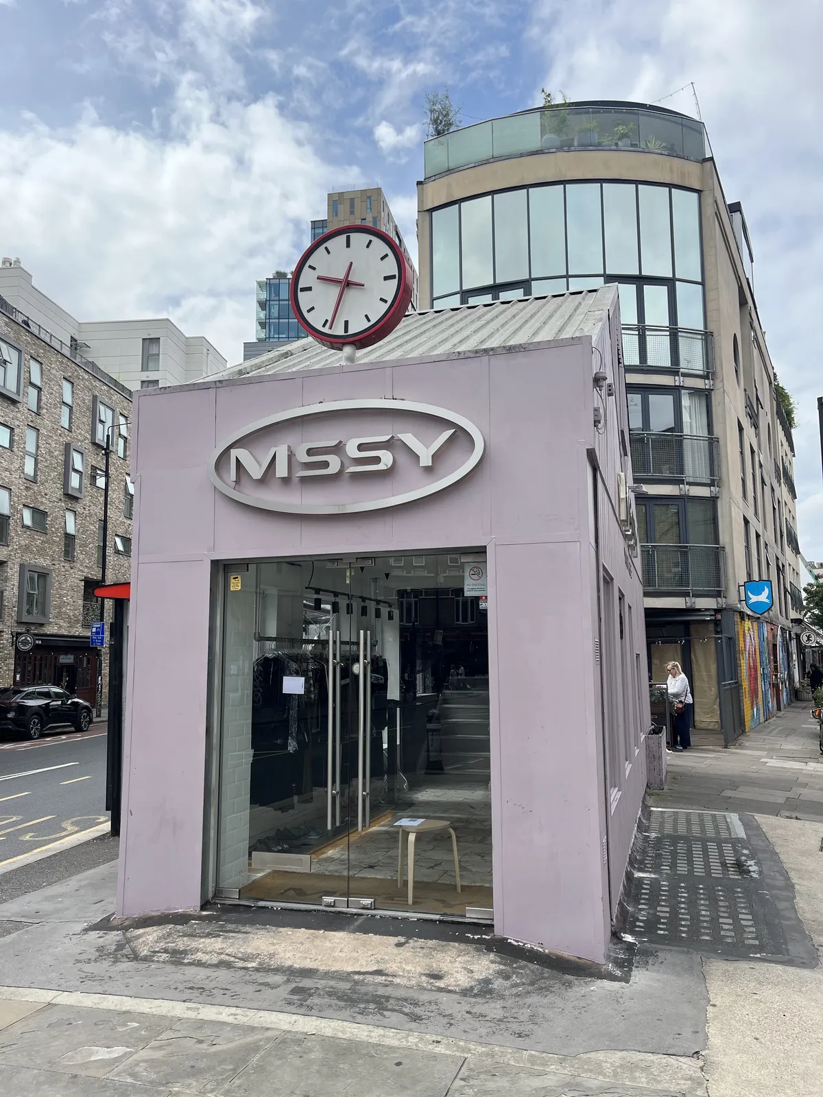 A clock on top of a small pink building in the middle of the street. It says MSSY on this building. The clock points at 34 minutes past nine.