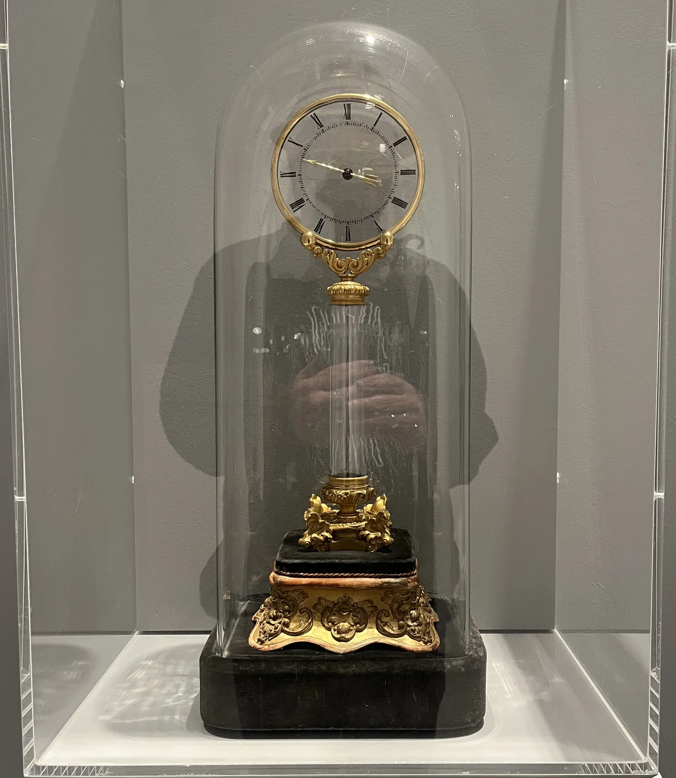 A clock in a glass bell, inside a glass showcase. The reflection of the photographer is visible, their head is perfectly aligned with the face of the clock. The clock seems magical: its single hand is attached to a glass plate, and there is absolutely no visible mechanism. Since it is a single hand, the time is not entirely precise. But if you look closely you can conclude that it points at approximately 35 minutes past nine.
