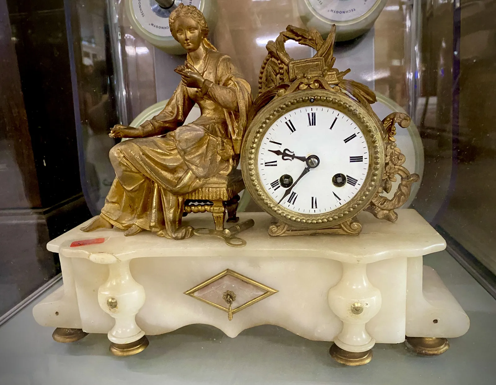 An elegant clock. It’s made from a golden statue of a sitting woman, leaning against a clock, on top of a white, marble pedestool.  The clock points at 36 minutes past 9.