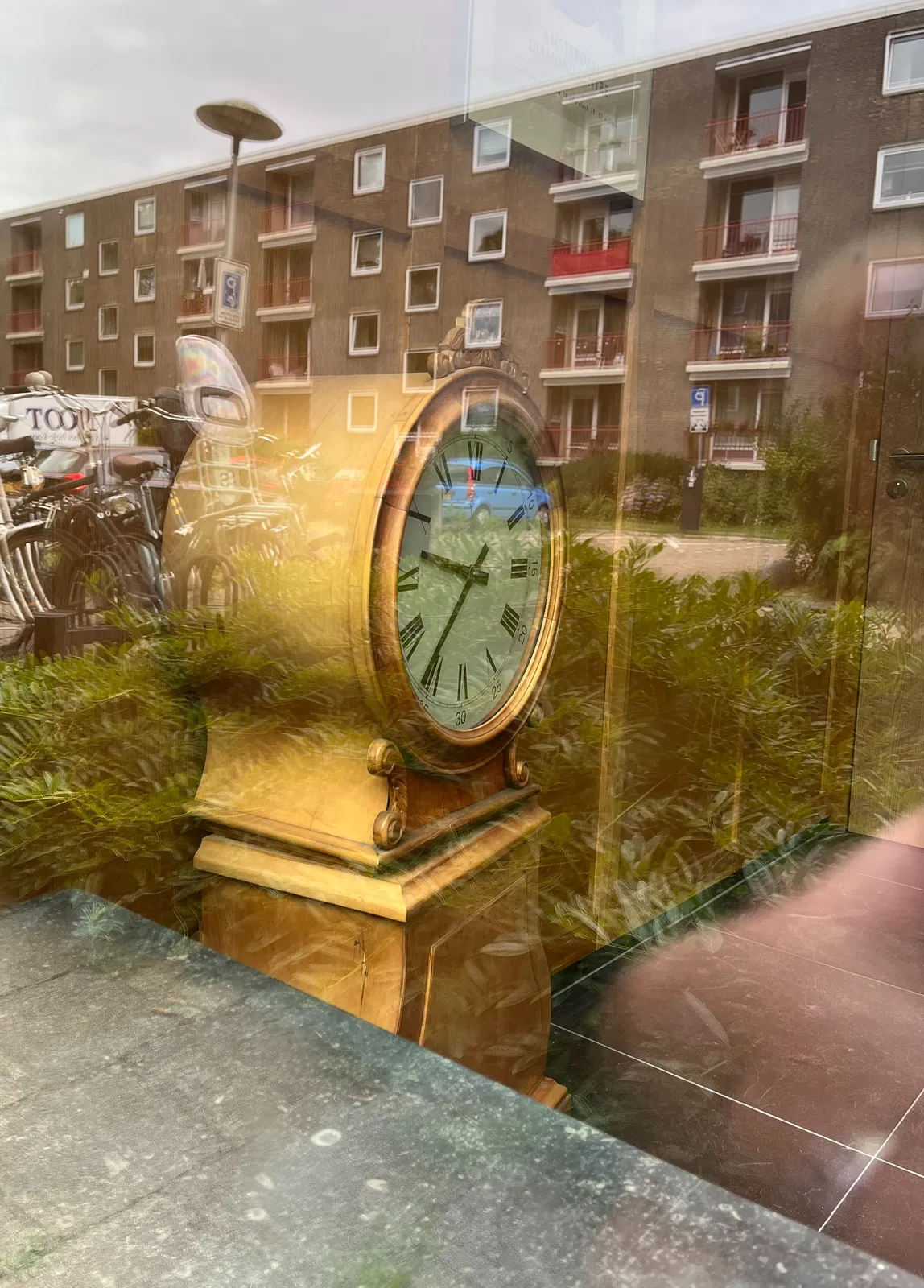 A appartment building can be seen in the reflection. Behind the window there’s a clock that points at approximately 36 past 9.