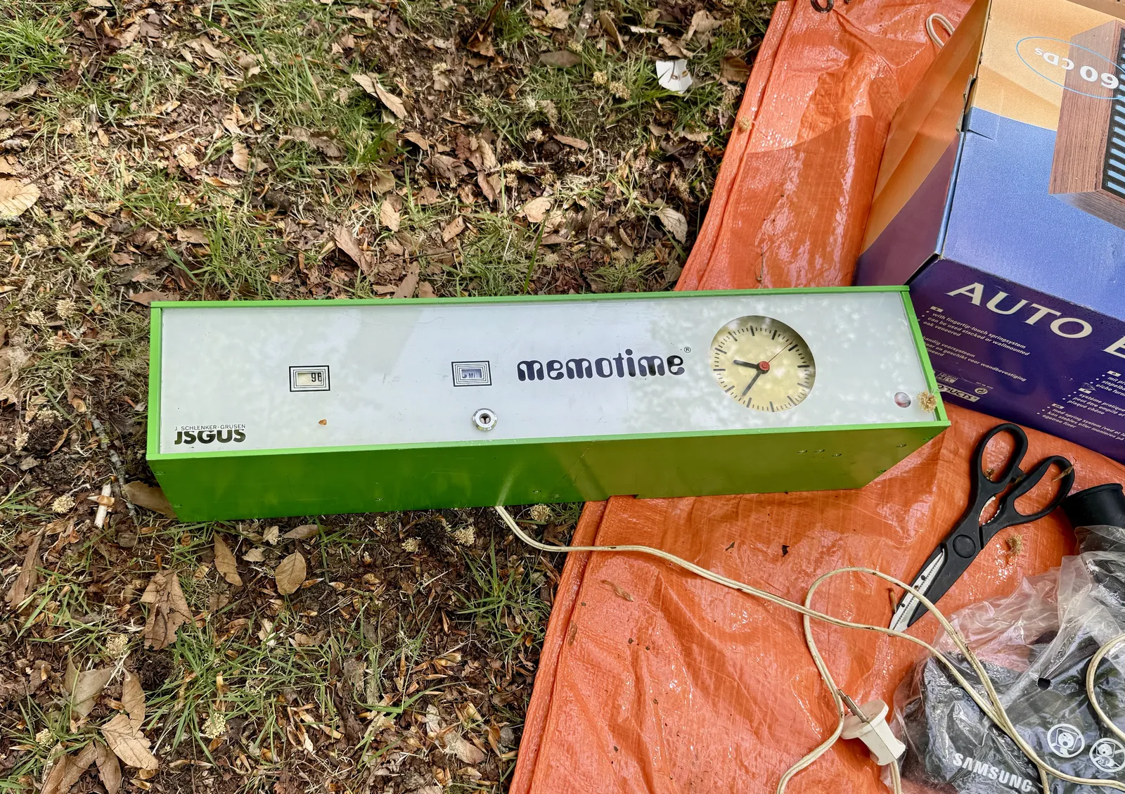 A wide, green, plastic, electronic device lies on the ground. It says Memotime in a seventies futuristic font, and J. Schlenker-Grusen is its maker. There are two little holes in the device. Behind the one on the left the number 96 is shown. Behind the one on the right it says mi. There’s also a clock on the thing, which points at 37 minutes past nine.  