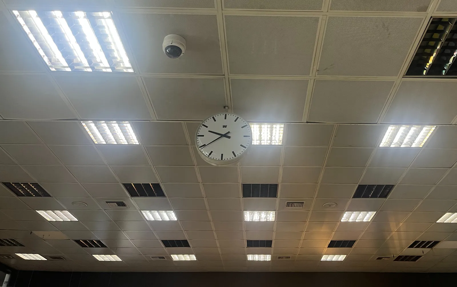 A clock hanging from a dropped ceiling. It points at 21 minutes to ten.
