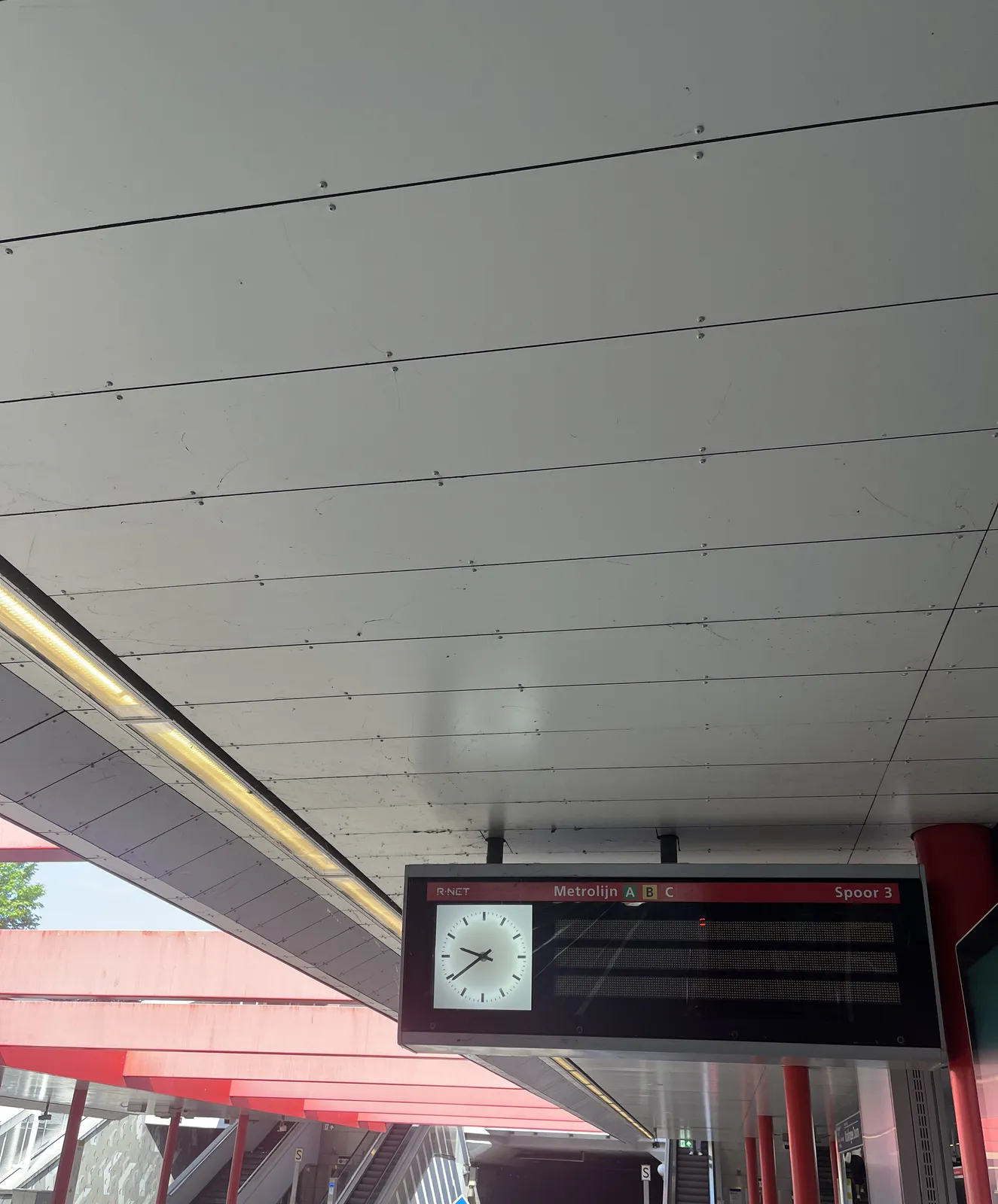 A digital display attached to the ceiling of a station. There are many red tones in this image. The clock on this display points at 39 minutes past 9.