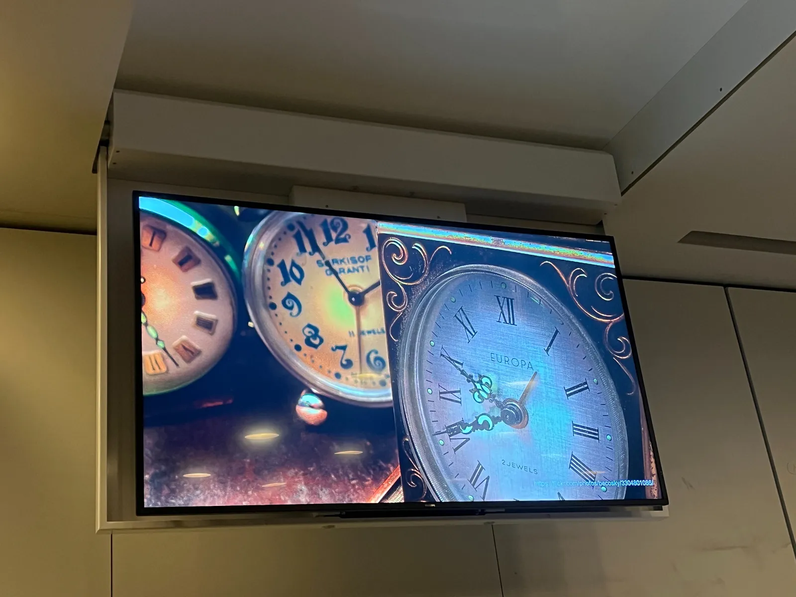 A picture of three clocks, shown on a screen which is lowered from the ceiling. On two of these clocks the time can be read. On one of these clocks it is 4 minutes to 2. And the other points at 20 minutes to 10. 