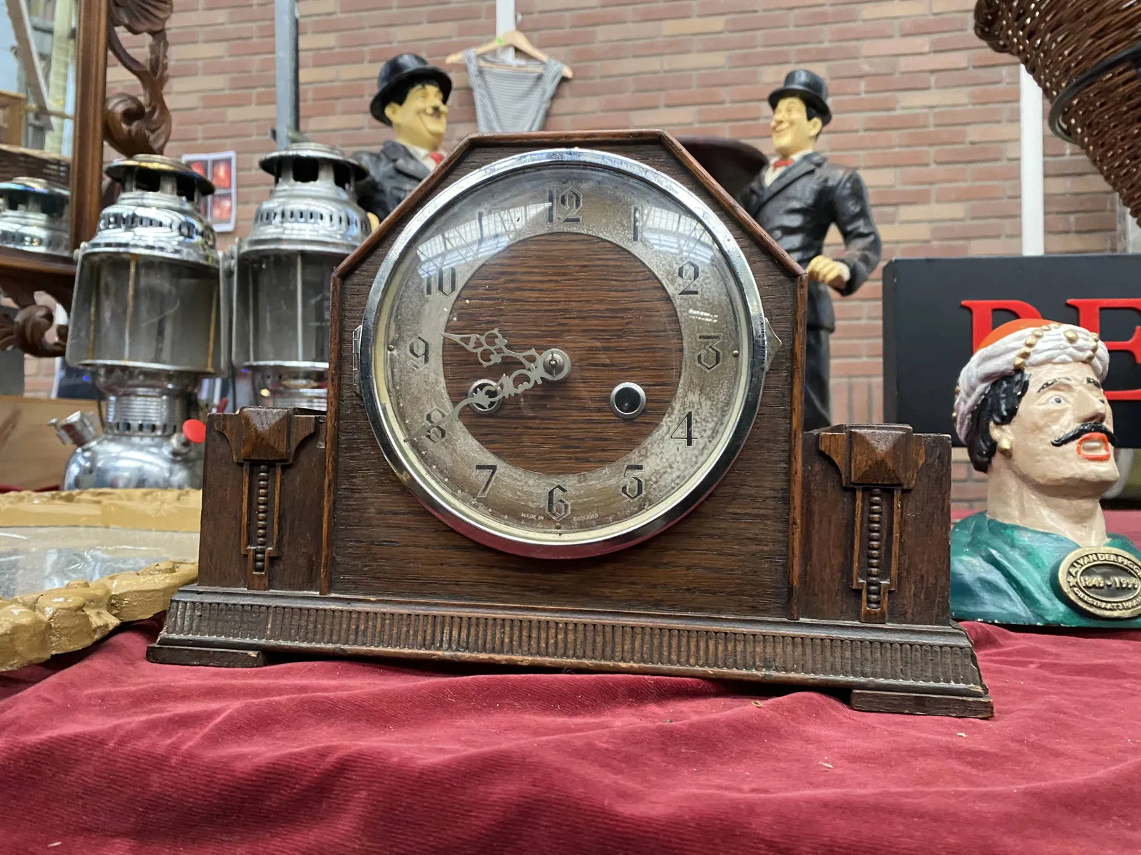A wooden clock stading among some old statues and some oils lamps on a flea market. It points at 20 minutes to 10.