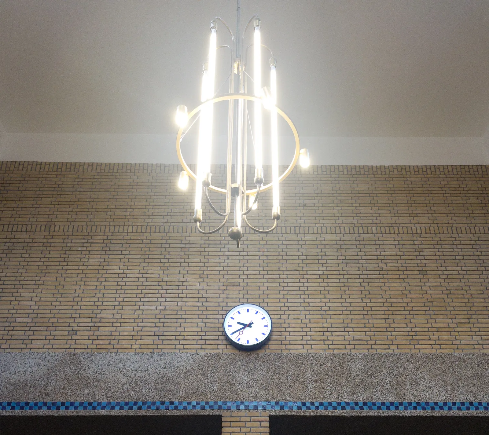 A chandelier made from neon tubes hangs from a high ceiling in front of a detailed brick wall. There’s a station clock on this wall which points at 20 minutes to ten.