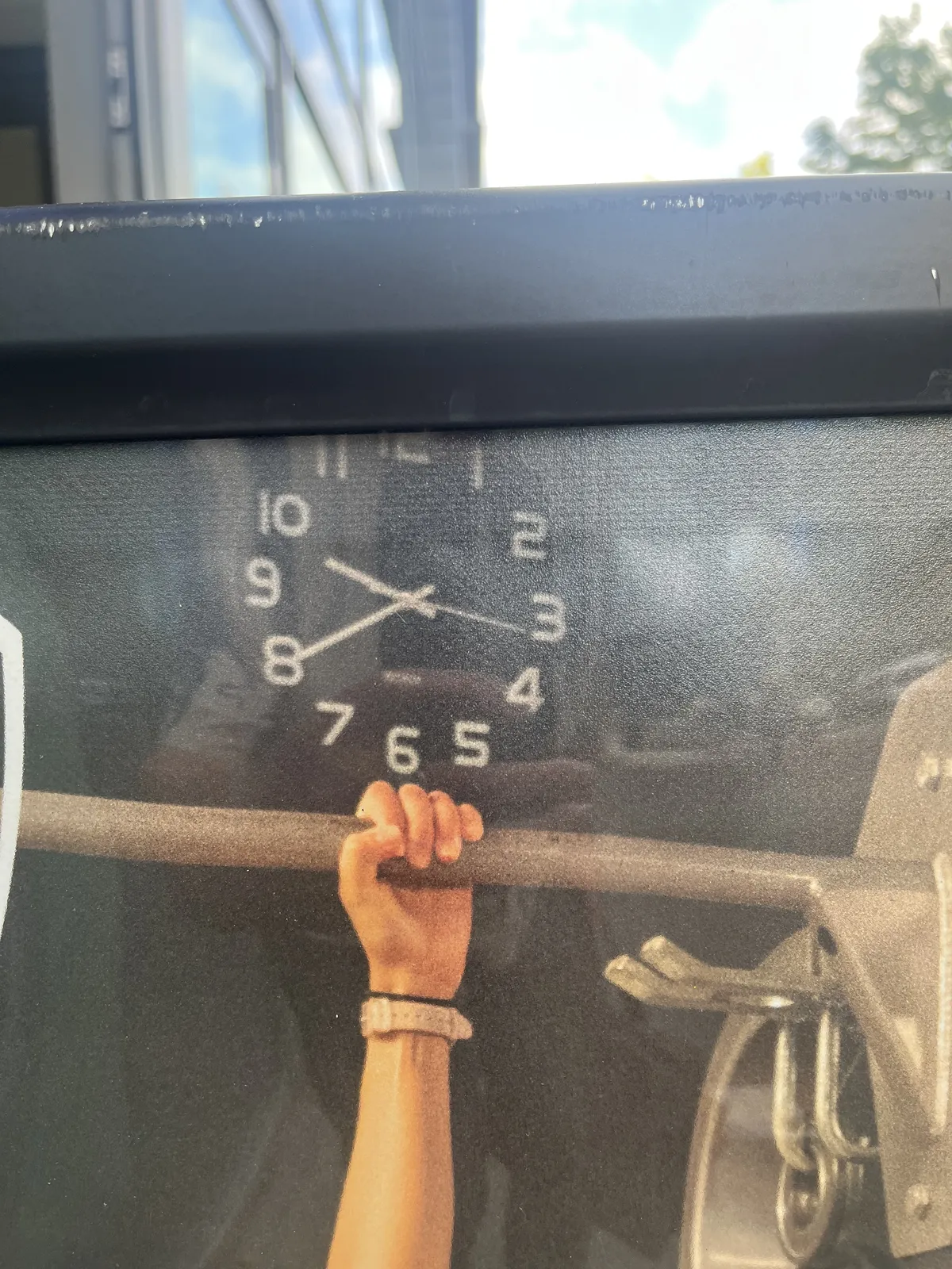 A detail of a billboard, outside. In the background some blurry trees and a blurry building can be seen. On the billboard you can see a hand, hanging from a pole. Behind the scene there’s a clock that points at 20 to ten.
