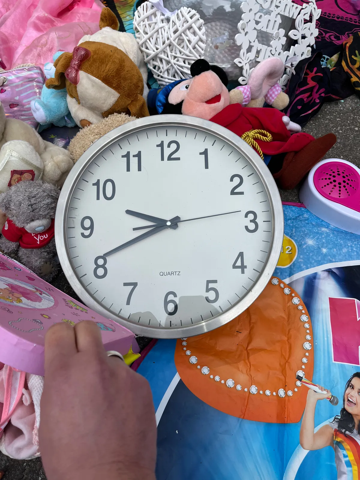 A clock lies on the ground, surrounded by cuddly toys and other toys. It points at 19 minutes to 10.