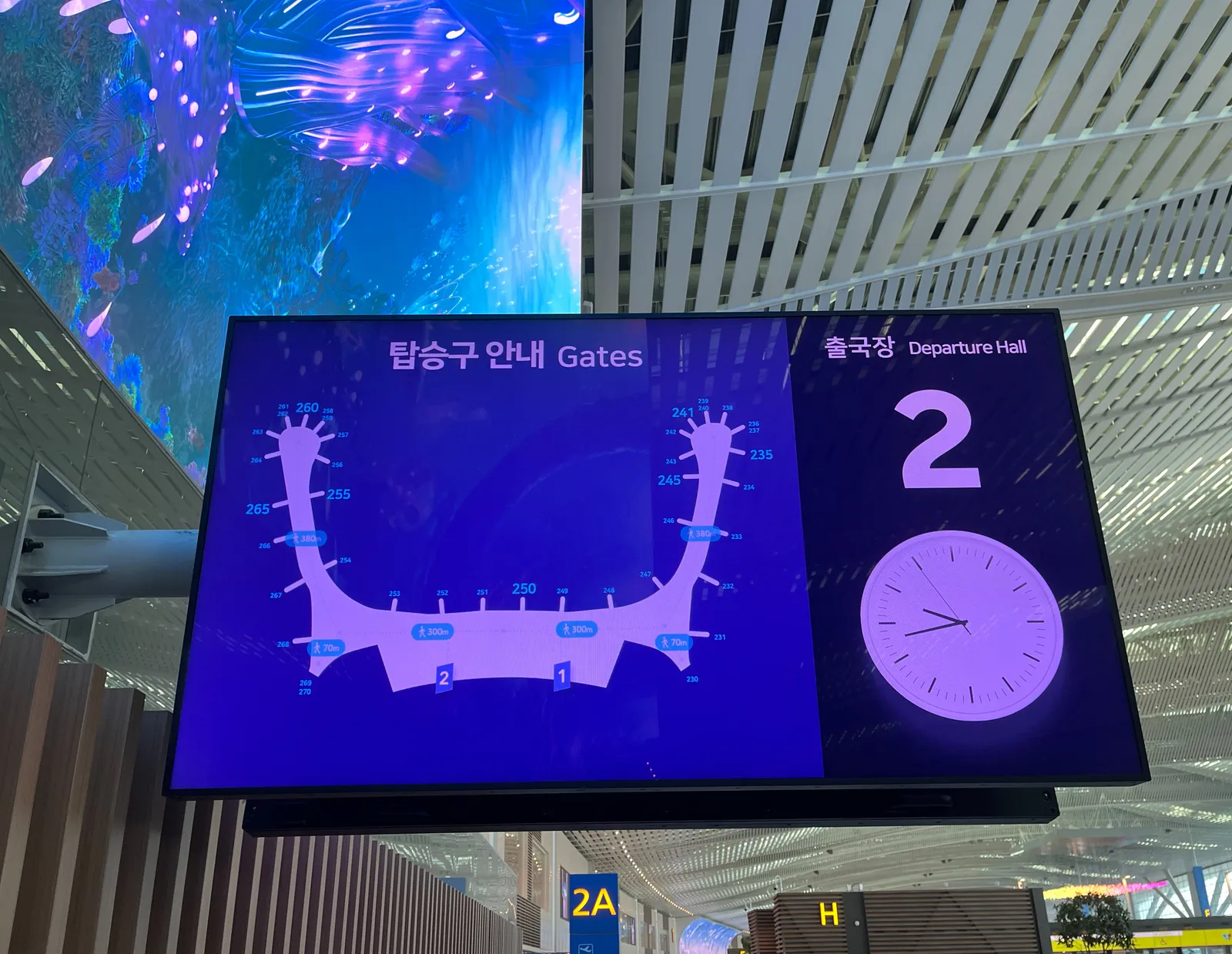 A sign with the plan of the airport. It hangs in an enormous terminal. There’s a gigantic curved TV screen hanging over this sign. And there’s also a clock. It points at 18 minutes to 10.