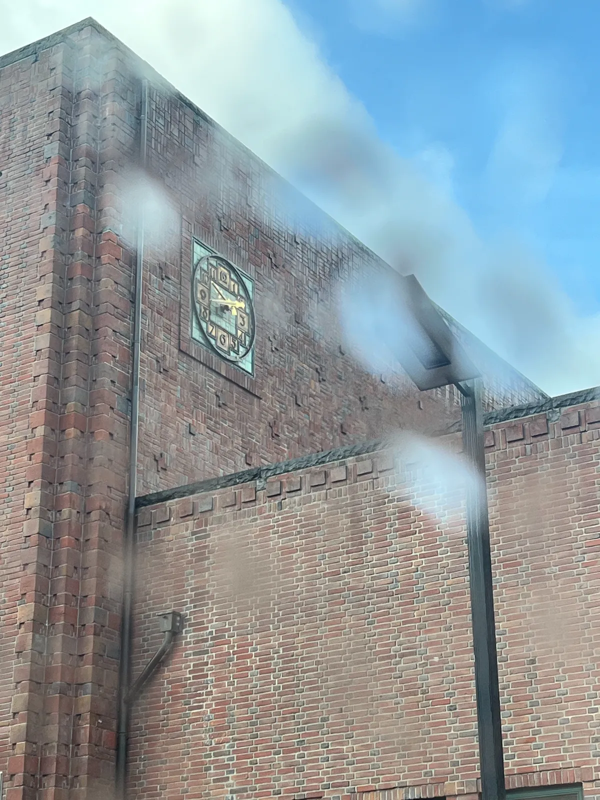 Looking through a window with rain drops on it at a monumental brick building with a cast iron clock cemented into its side. It points at 17 minutes to 10.