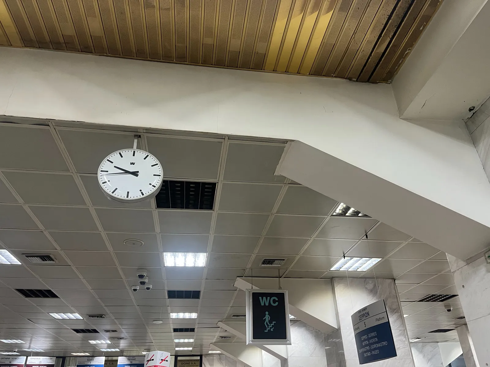 A clock hanging from a dropped ceiling. There’s also a sign pointing at the toilets. The clock points at 16 minutes to ten.
