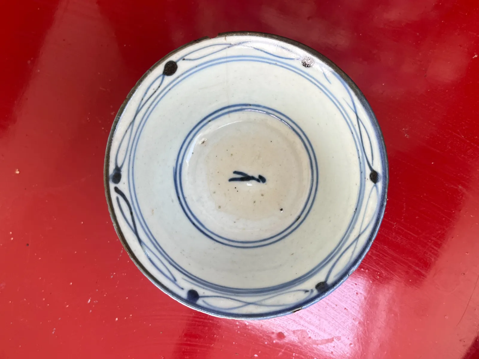 Looking down at a cup, straight from above. It stands on a red table. The cup is white and it has blue ornaments. In the center a non-latin letter is painted. This letter can also be seen as the hands of a clock, and if you do so, it points at a quarter to ten.
