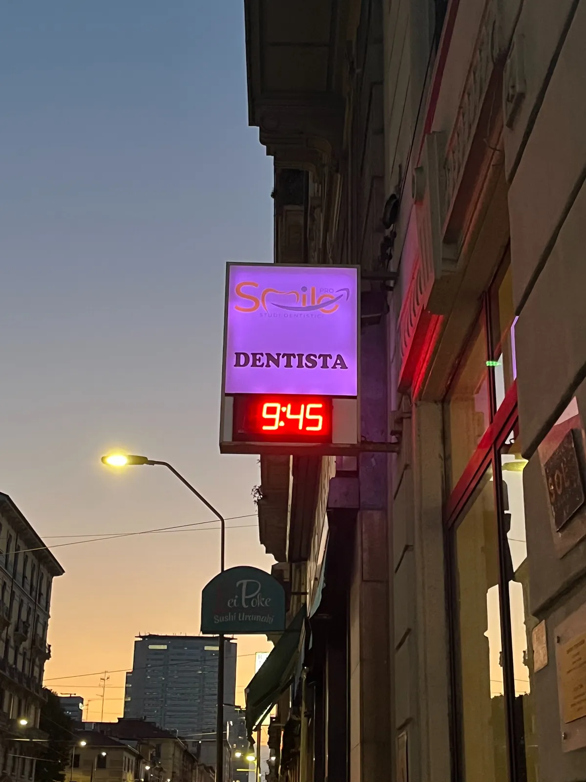 A sign attached to a building, indicating that Smile Dentista is right here. There’s also a digital clock in this sign which says the time is 09:45.