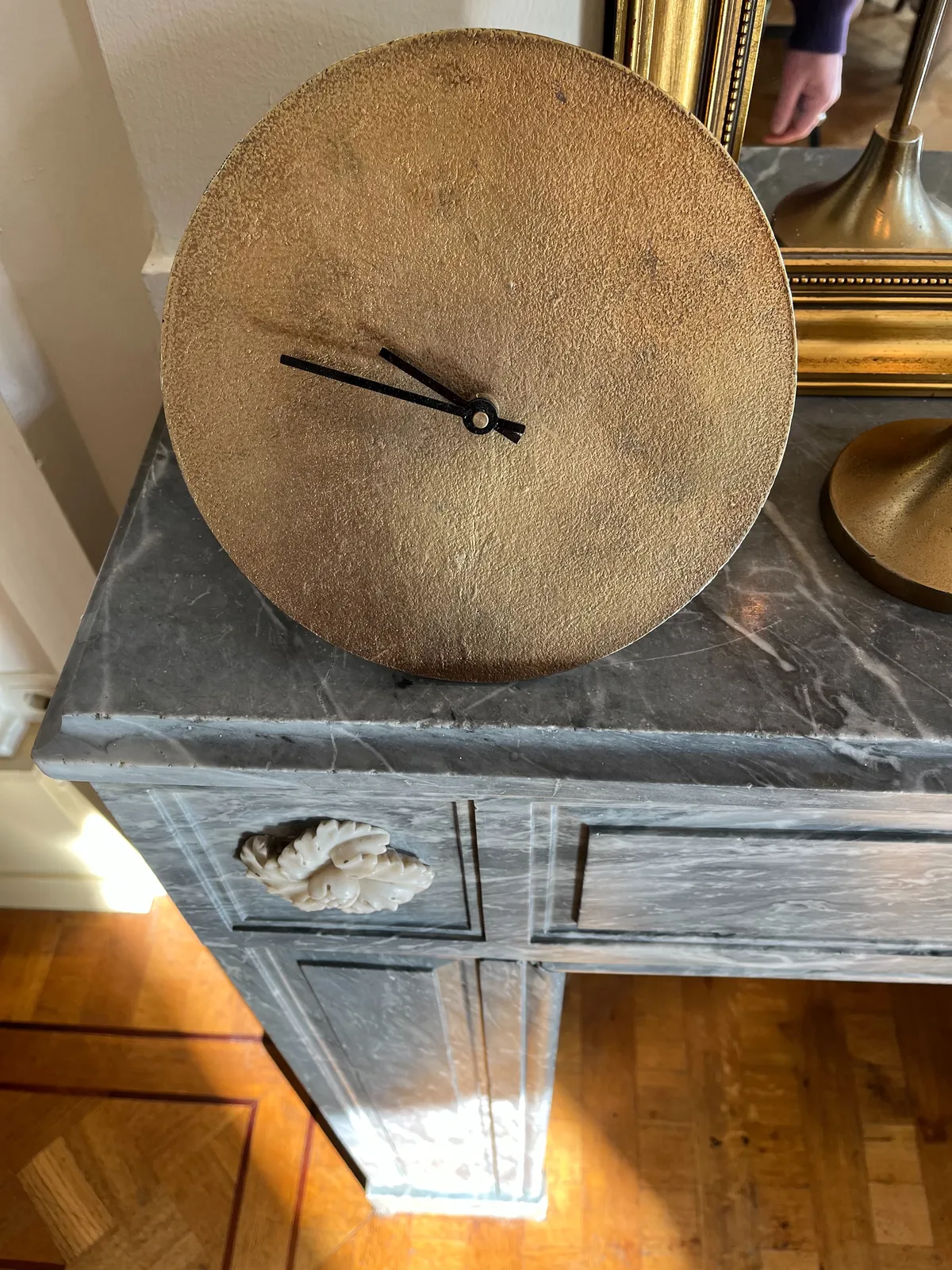 Looking down at a rather minimalist brass clock that stands on a mantel. The clock points at approximately 13 minutes to 10.