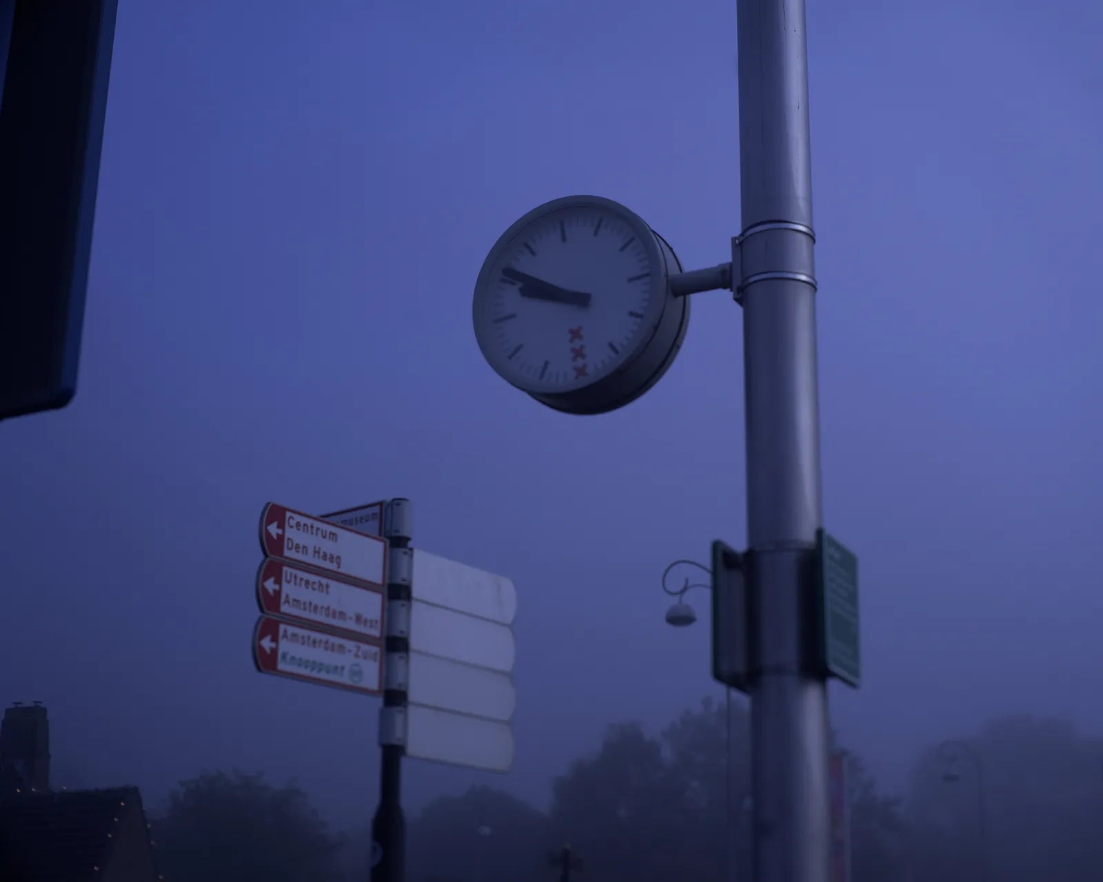 A signpost pointing in the direction of Centre, The Hague, Utrecht, Amsterdam West, and Amsterdam South. Next to it is a clock which points at 9 minutes to 10. 