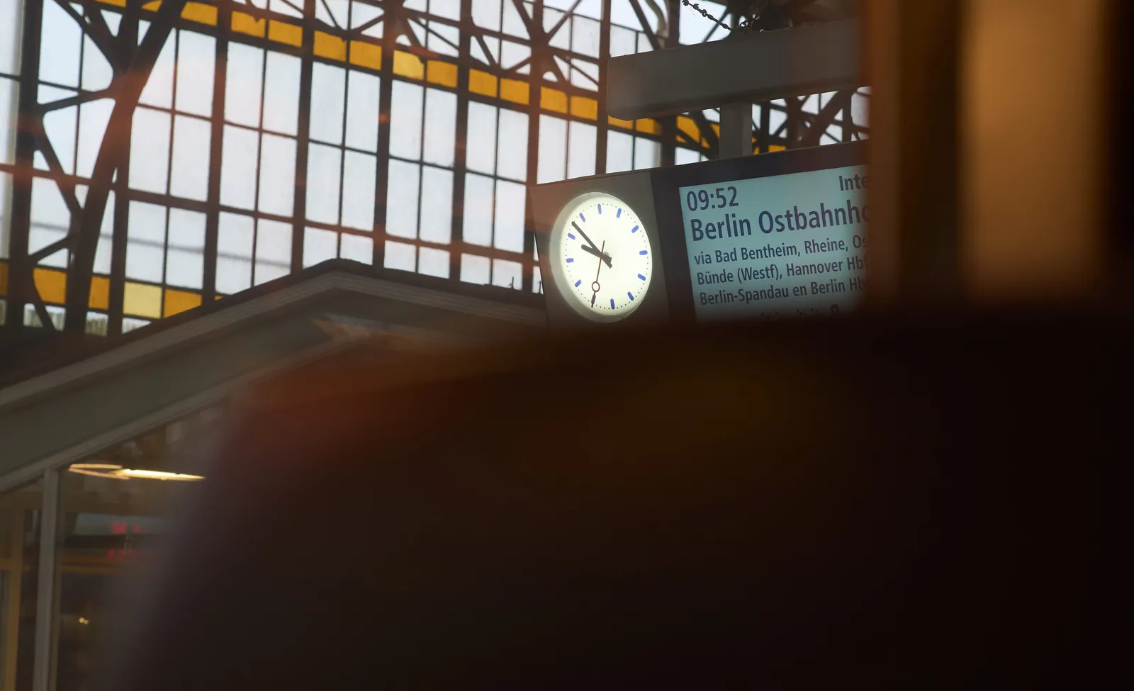 Peeking over the chairs in front of you, from inside a train, to the display that says that the train to Berlin Ostbahnhof will leave at 8 minutes to 10. The clock next to the display points at exactly 8 minutes to 10.