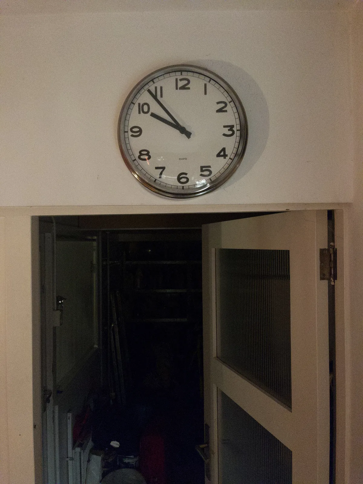 Looking through a half open door into a dark storage room. Above the door there’s a clock that points at 7 minutes to 10.