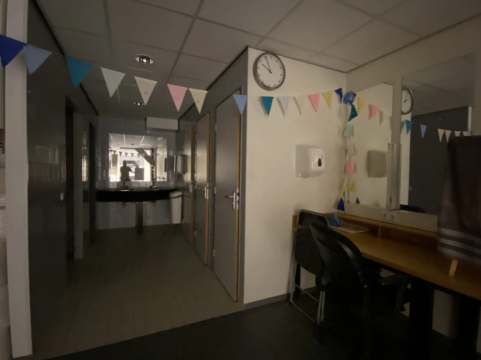 There’s a colourful paper chain in this dressing room, which doesn’t really succeed in making it look like a party. In the background, in the mirror, the photographer can be seen. On a wall there’s a clock which points at 4 minutes to 10.