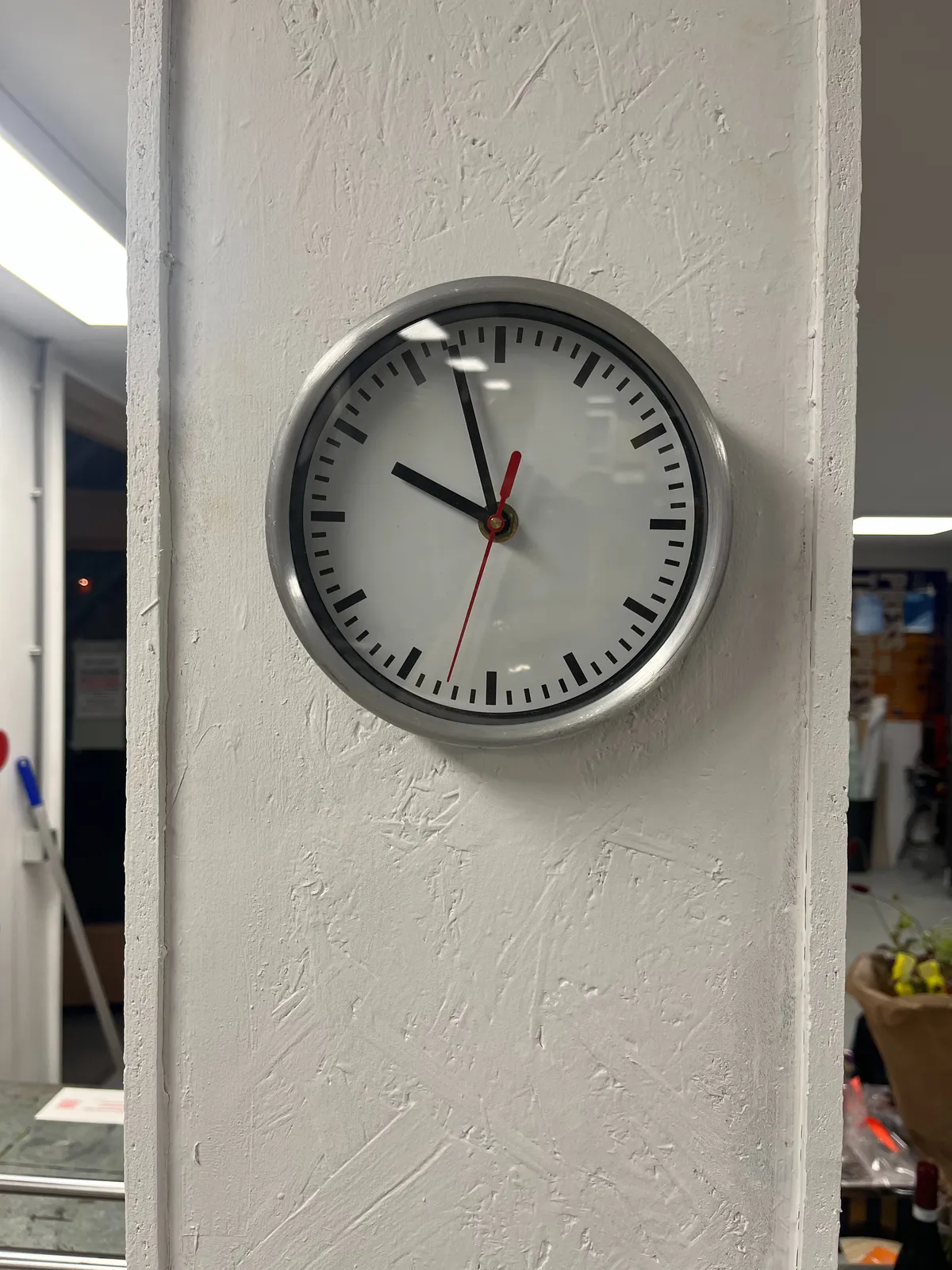 A clock on a pillar in a rather large room. It points at 3 minutes to 10.