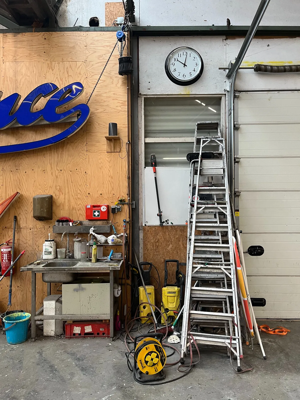 A wall in a workshop. There are ladders, a sink, and a clock. It points at 2 minutes past 10.